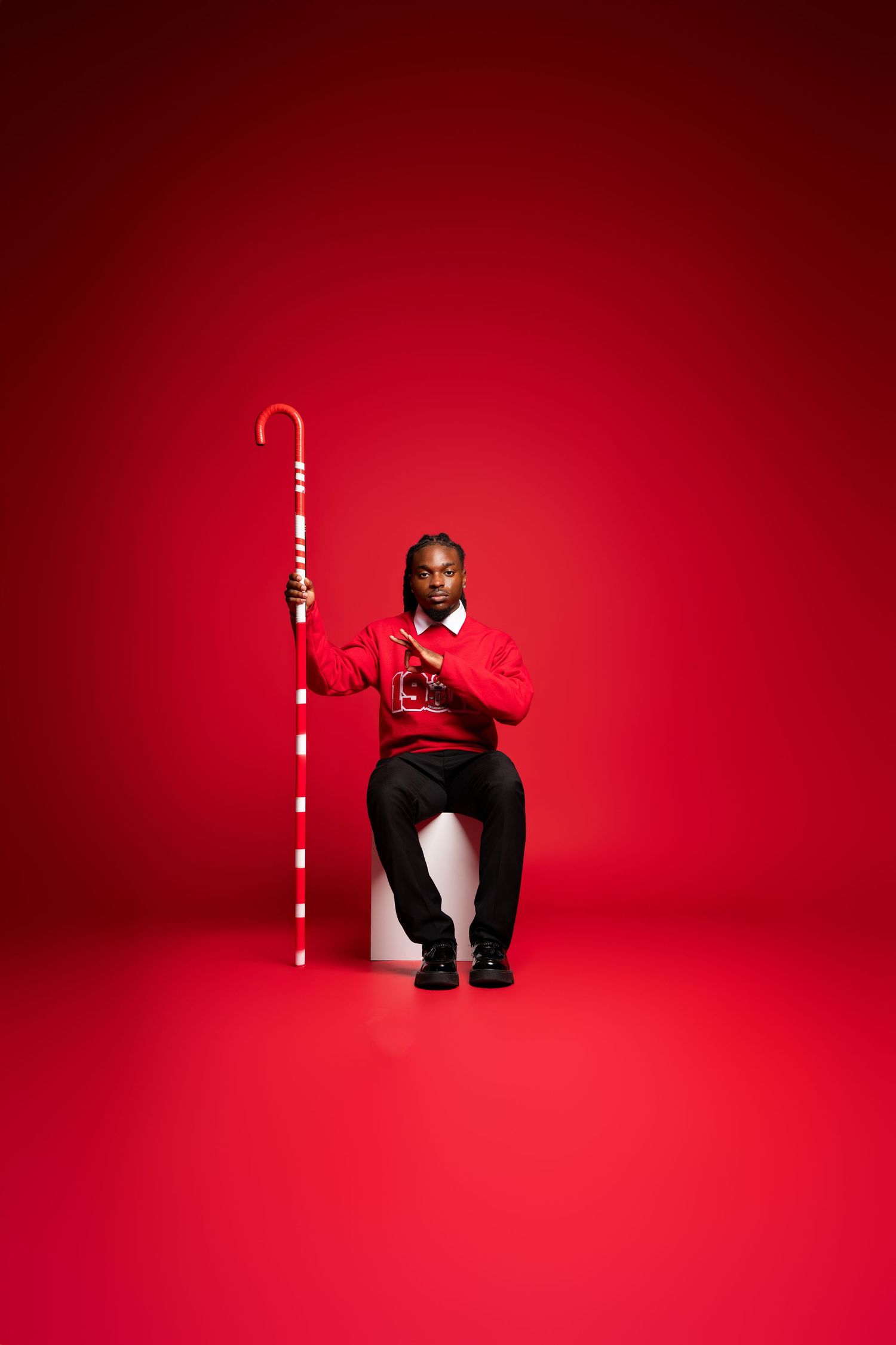 Man in red sweater sitting on white cube holding oversized candy cane against vibrant red background.