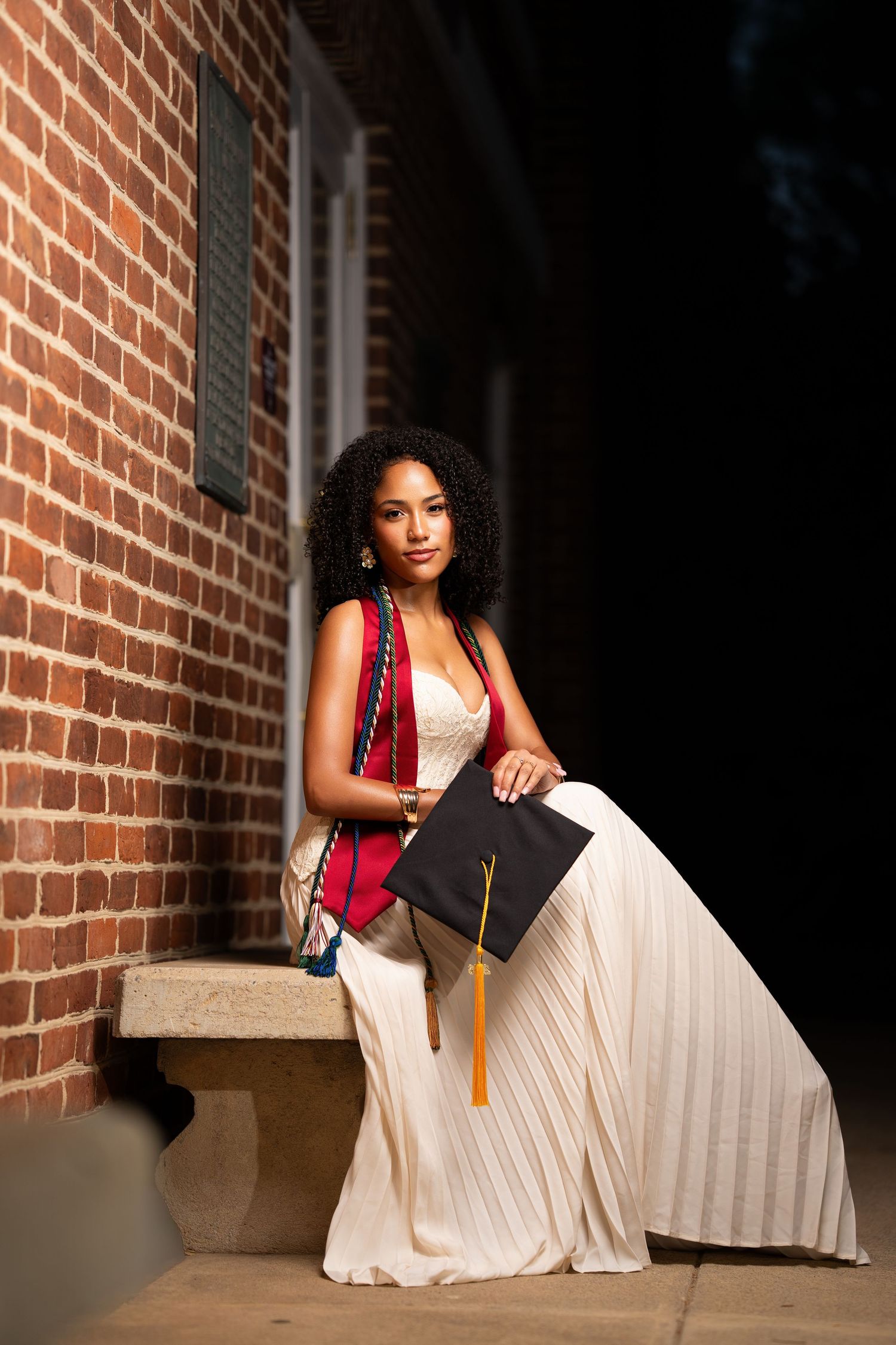 Young woman in white gown holding graduation cap beside brick wall, celebrating academic achievement.