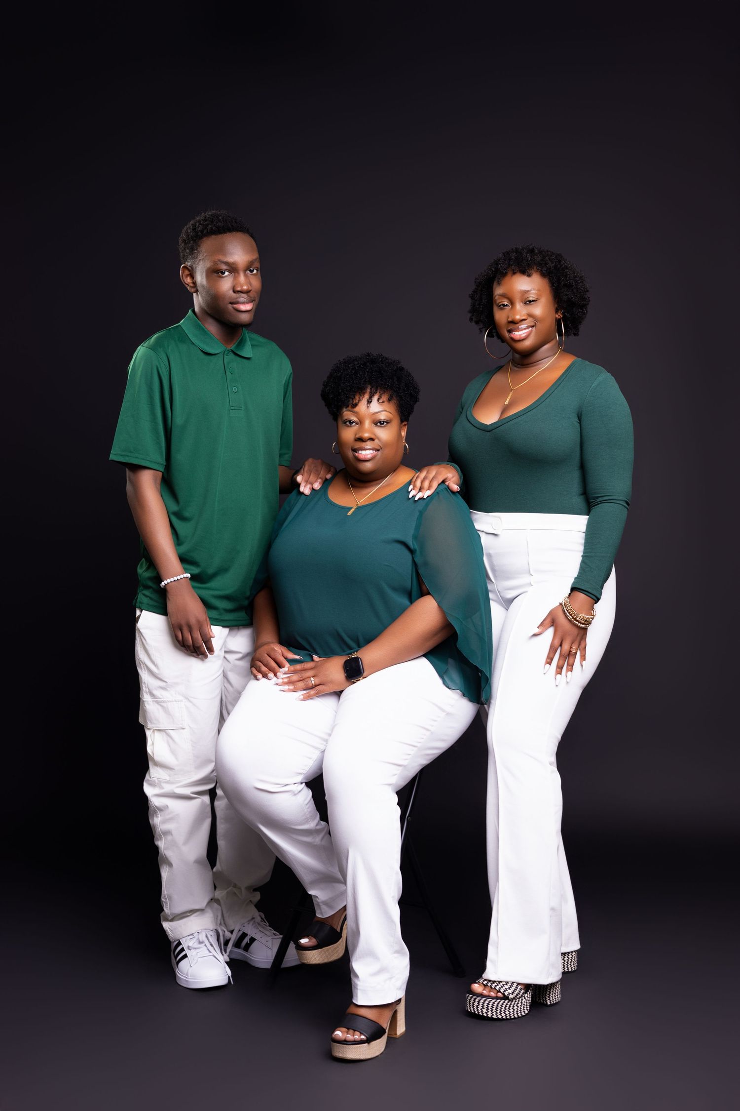 Three people in matching green tops and white pants pose for a family portrait against a dark backdrop.