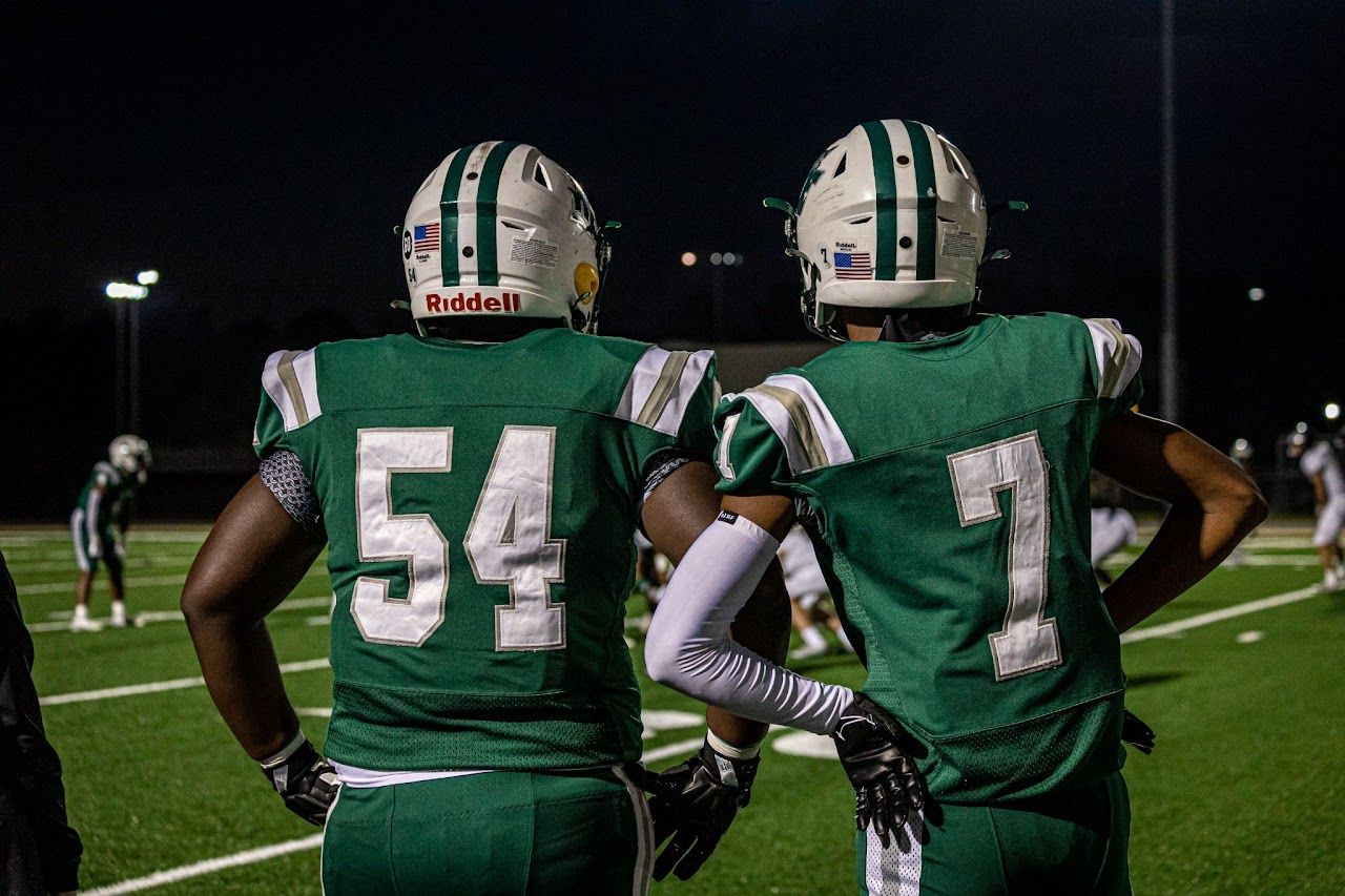 Two football players wearing green and white uniforms with numbers 54 and 7 stand on a field at night.  DFHS