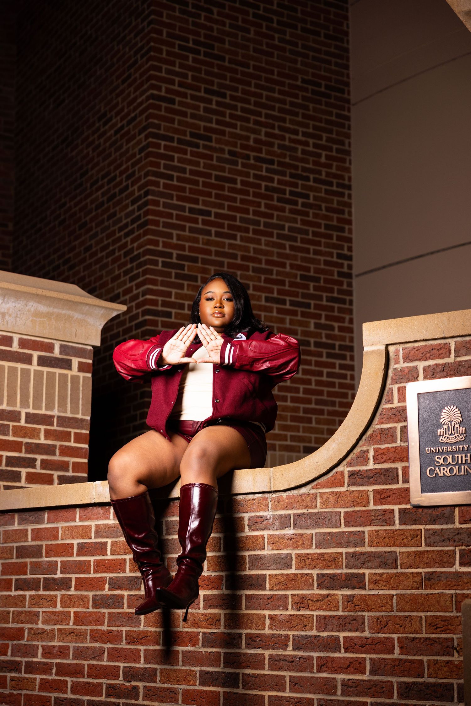 Graduate in red outfit posing on brick wall at dusk.
