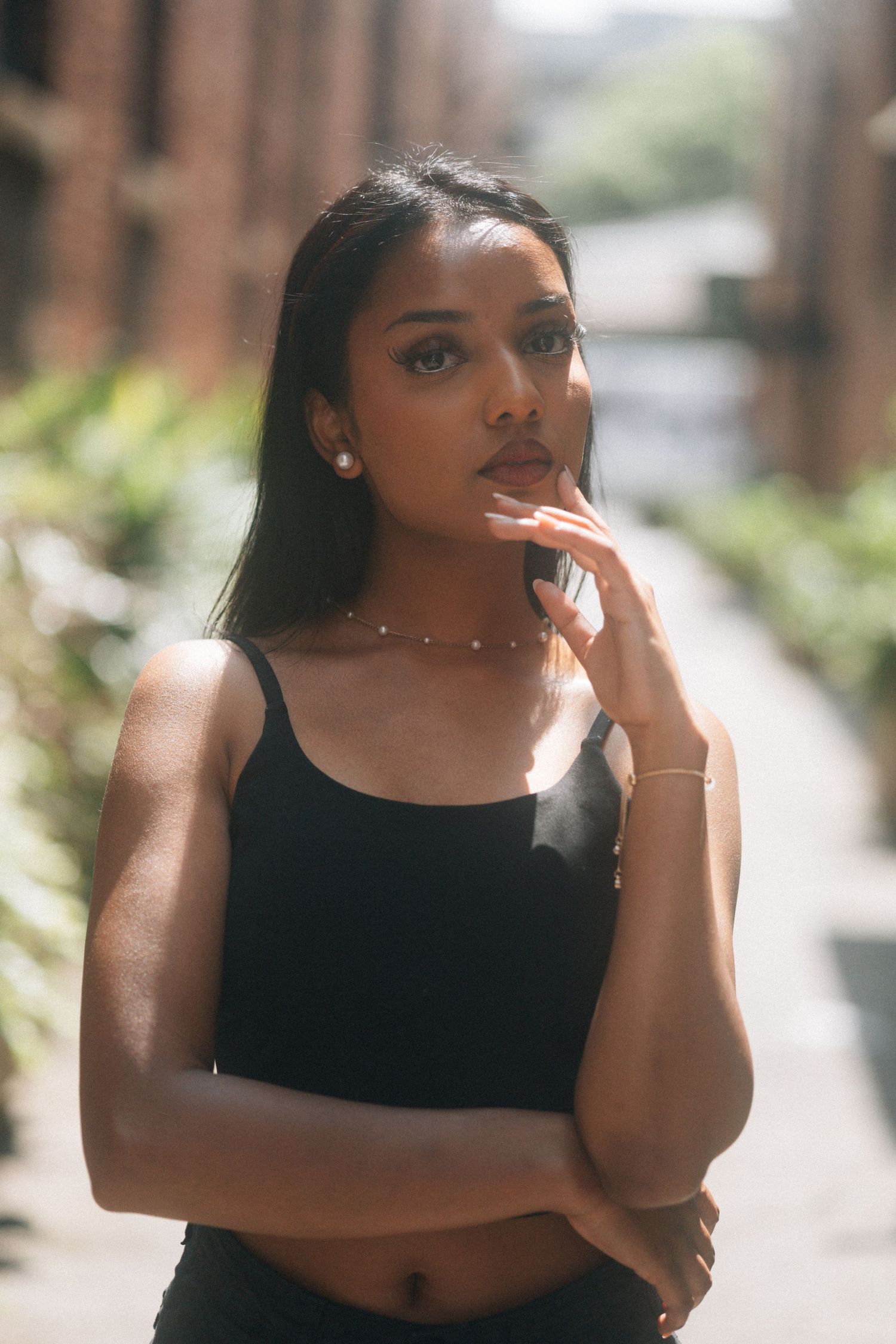 A thoughtful pose outdoors in natural lighting wearing a black tank top with delicate jewelry and long straight hair.