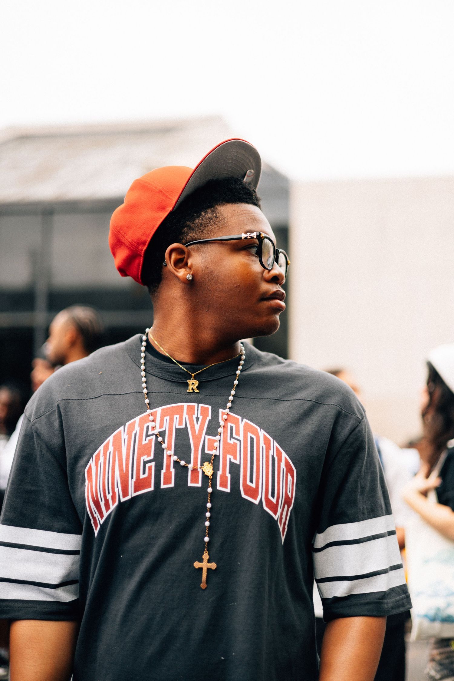 Person wearing a black varsity-style t-shirt with orange text and a cross necklace against an outdoor background.
