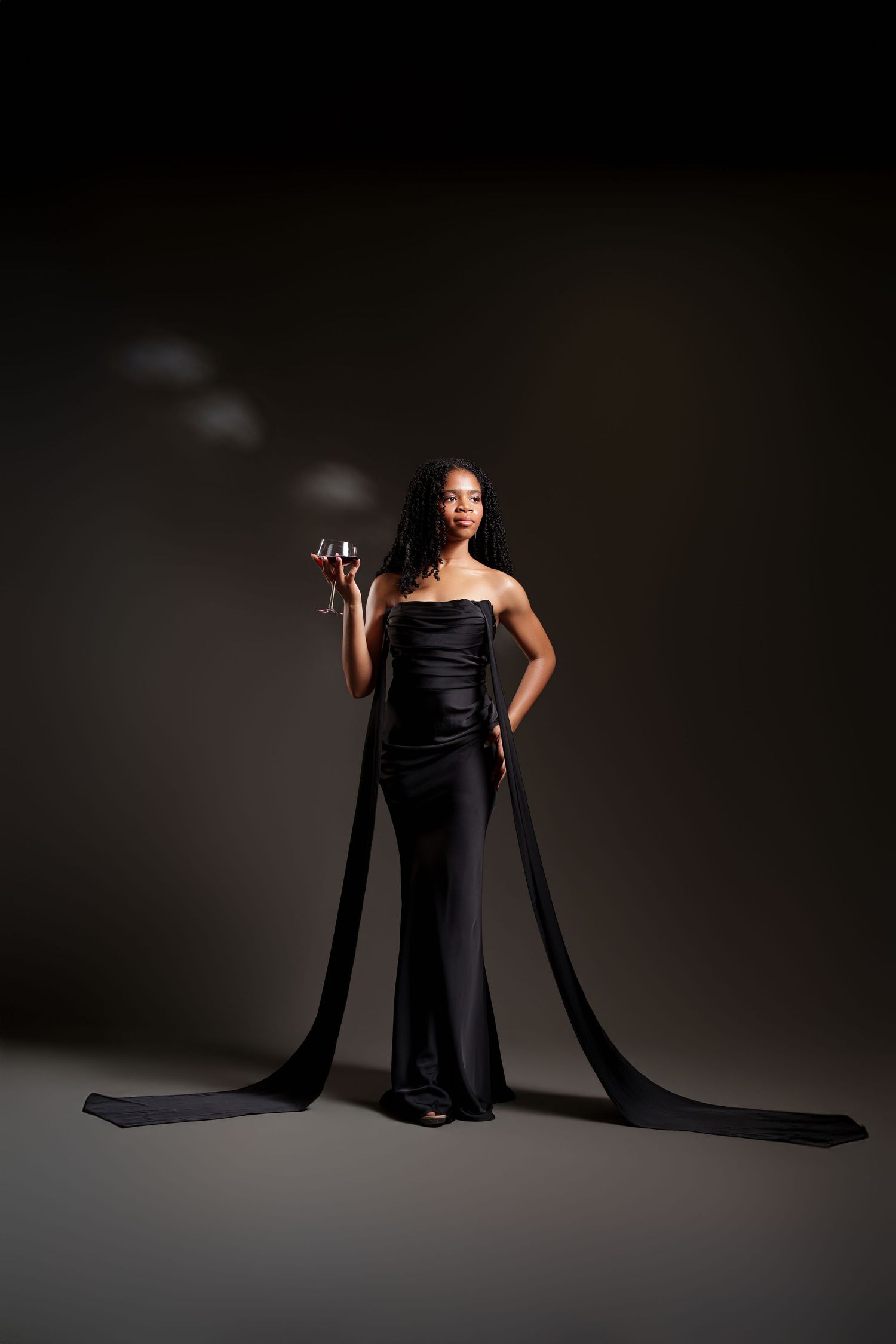 Elegant figure in black evening gown with dramatic flowing fabric panels holds wine glass against dark background.