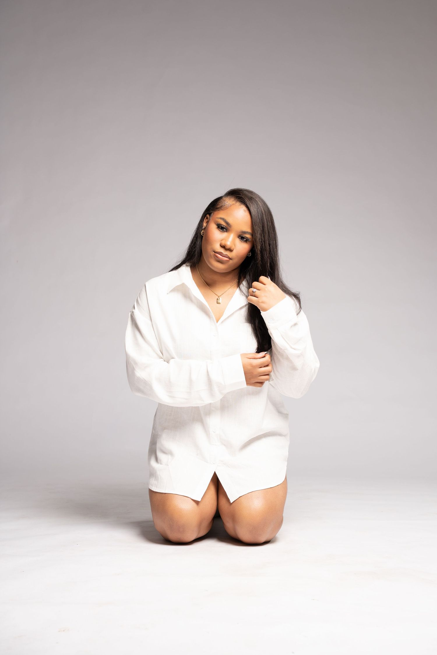 A person poses on their knees wearing an oversized white button-down shirt against a light gray studio background.