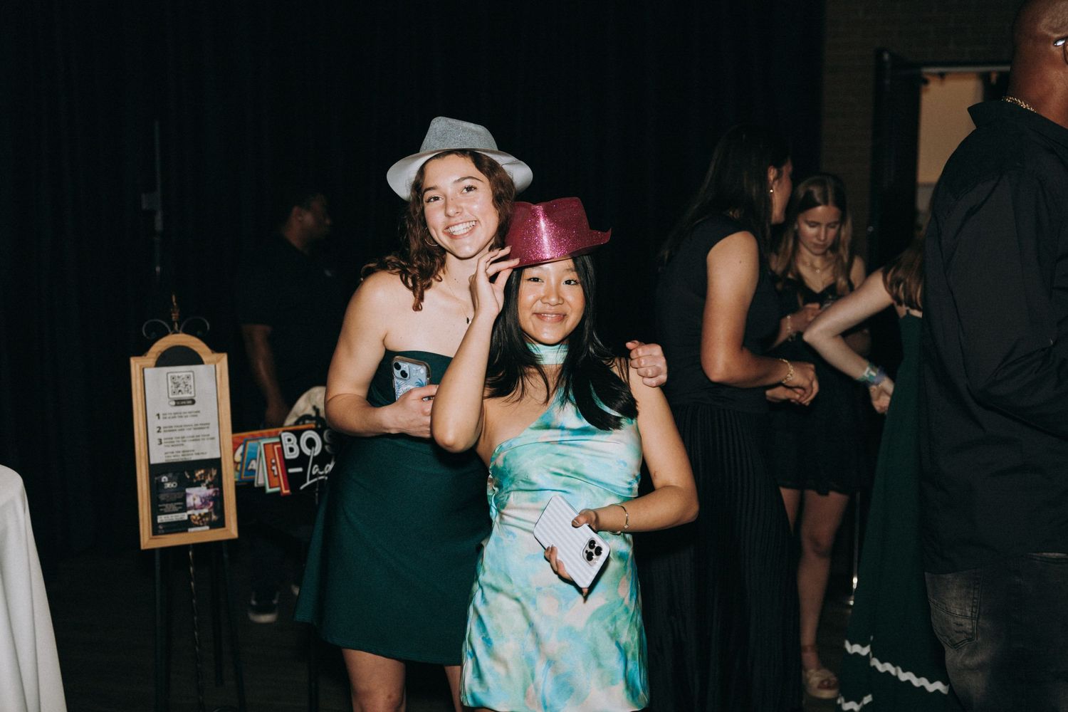 Two young women posing and smiling at a party, wearing fun hats, with guests dancing in the background.