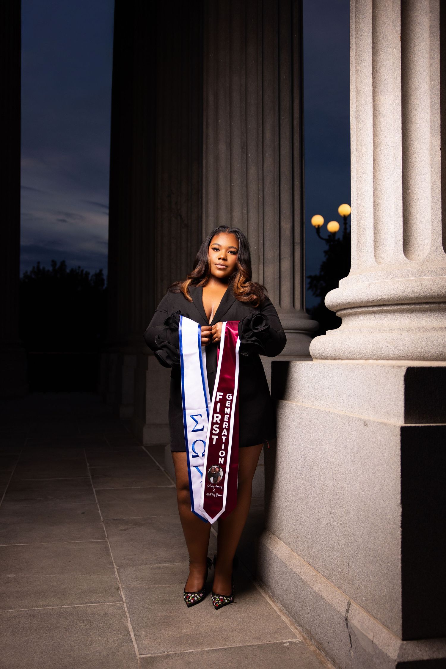 A young woman in a black dress poses with graduation stoles between large stone columns at dusk.