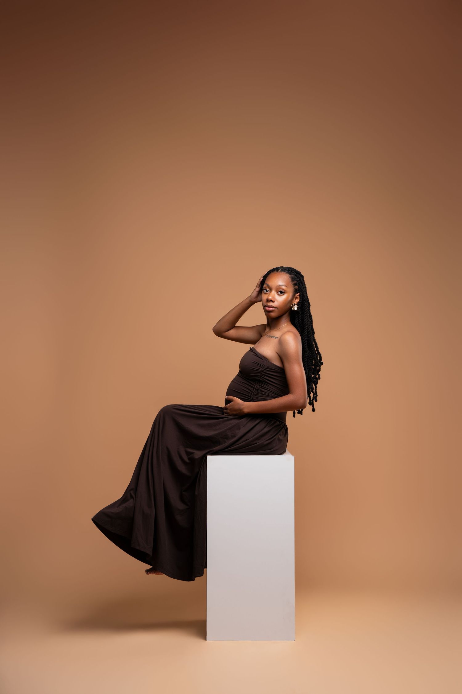 A young Black woman in a dark brown strapless gown sits gracefully on a white pedestal against a beige background.