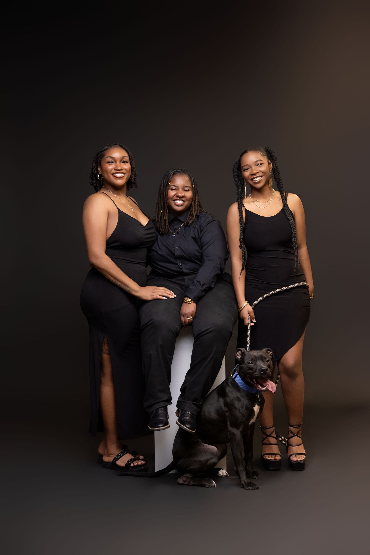 Three people in black outfits pose with a dark dog against a dark background in a studio setting.