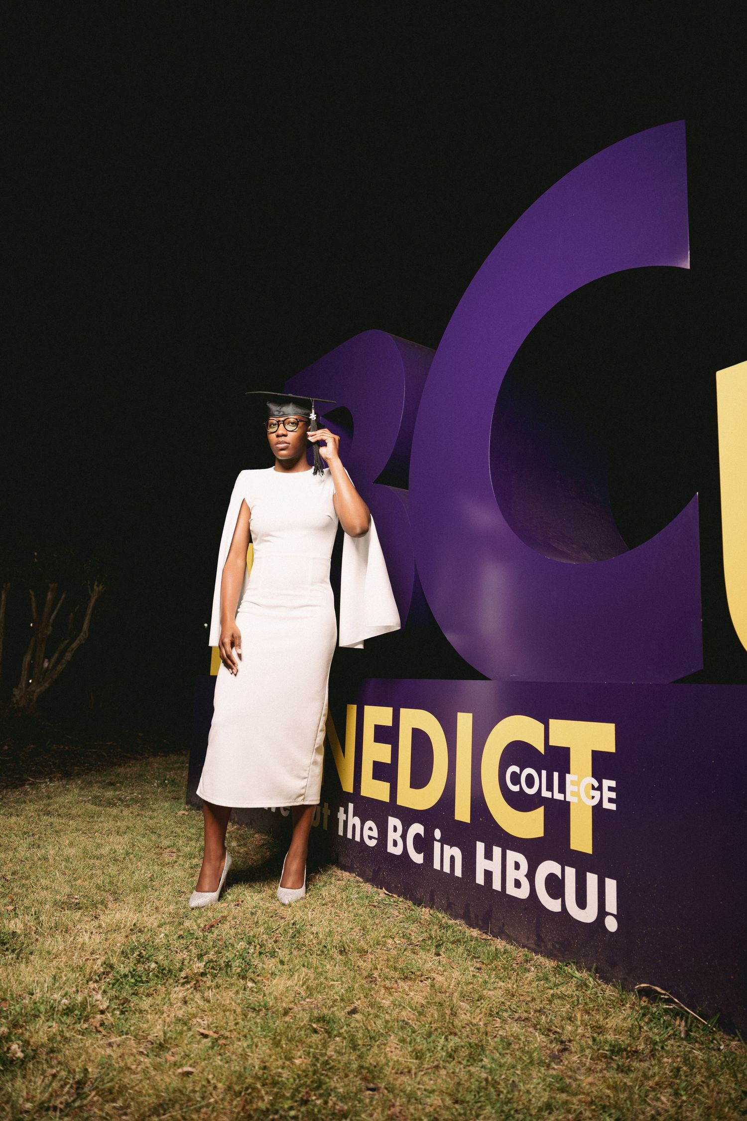 Graduate in white dress and cap poses in front of purple college sign outdoors.