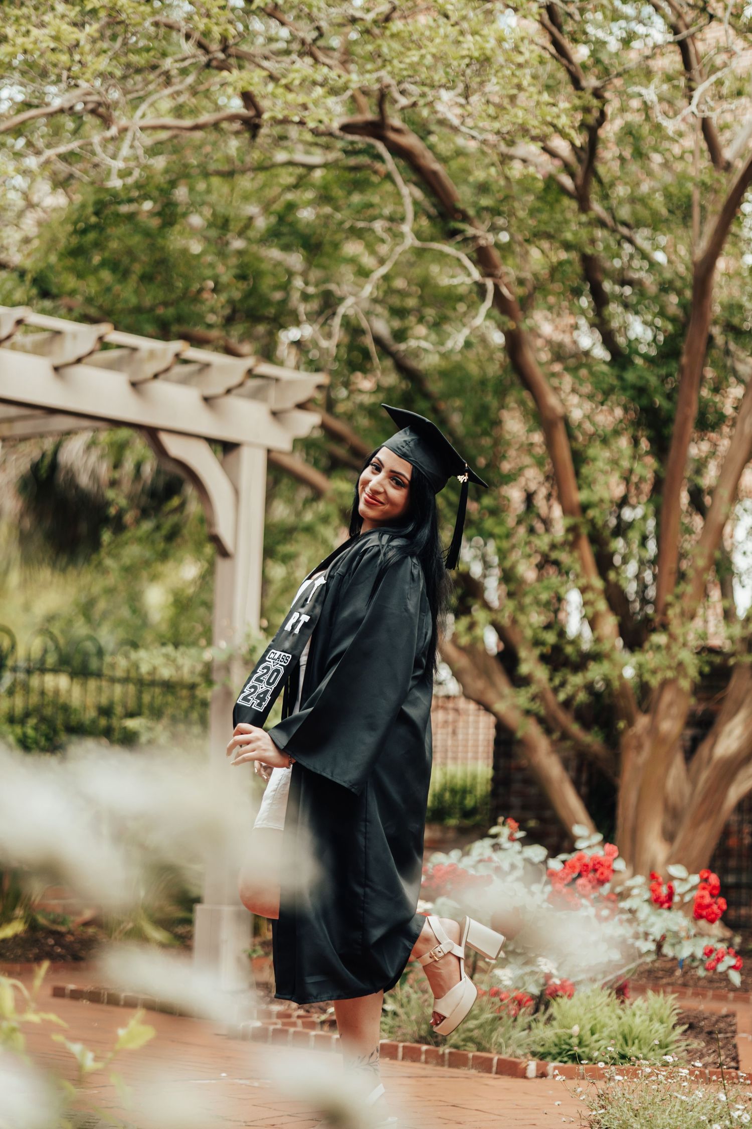 Graduate in green academic regalia poses in a garden setting with flowering trees and a wooden pergola.