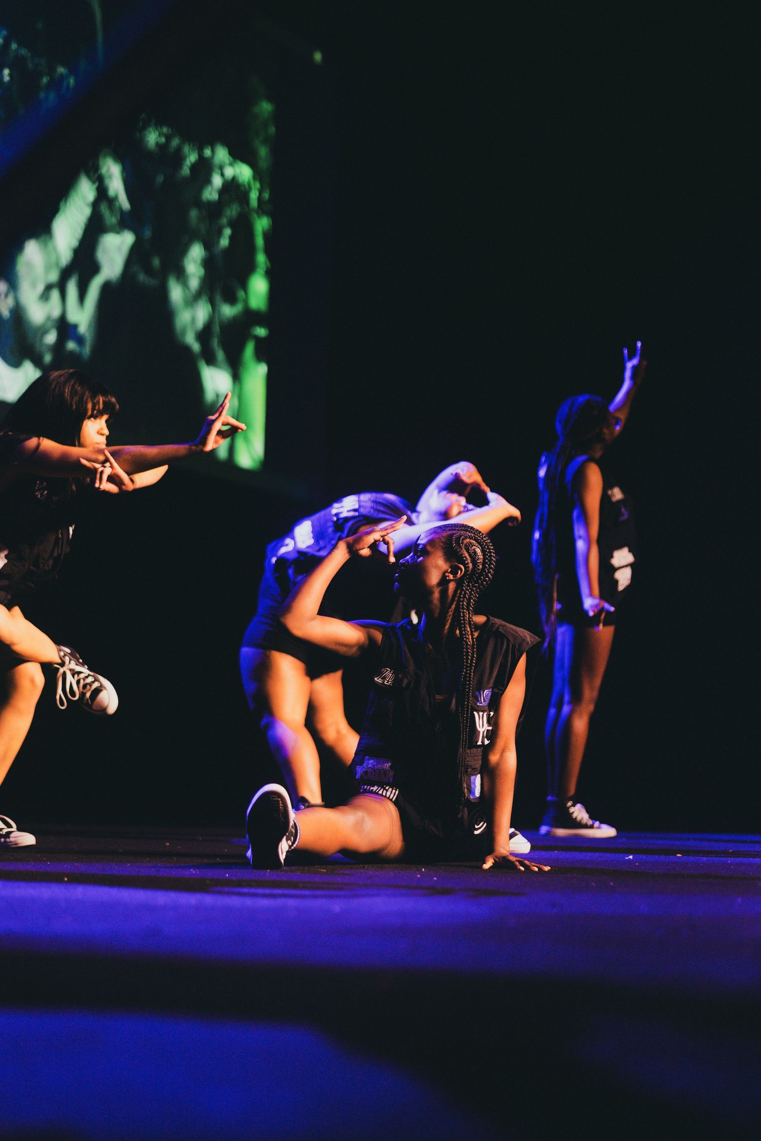 zetas in blue costumes perform a synchronized floor routine under dramatic stage lighting on a dark stage.