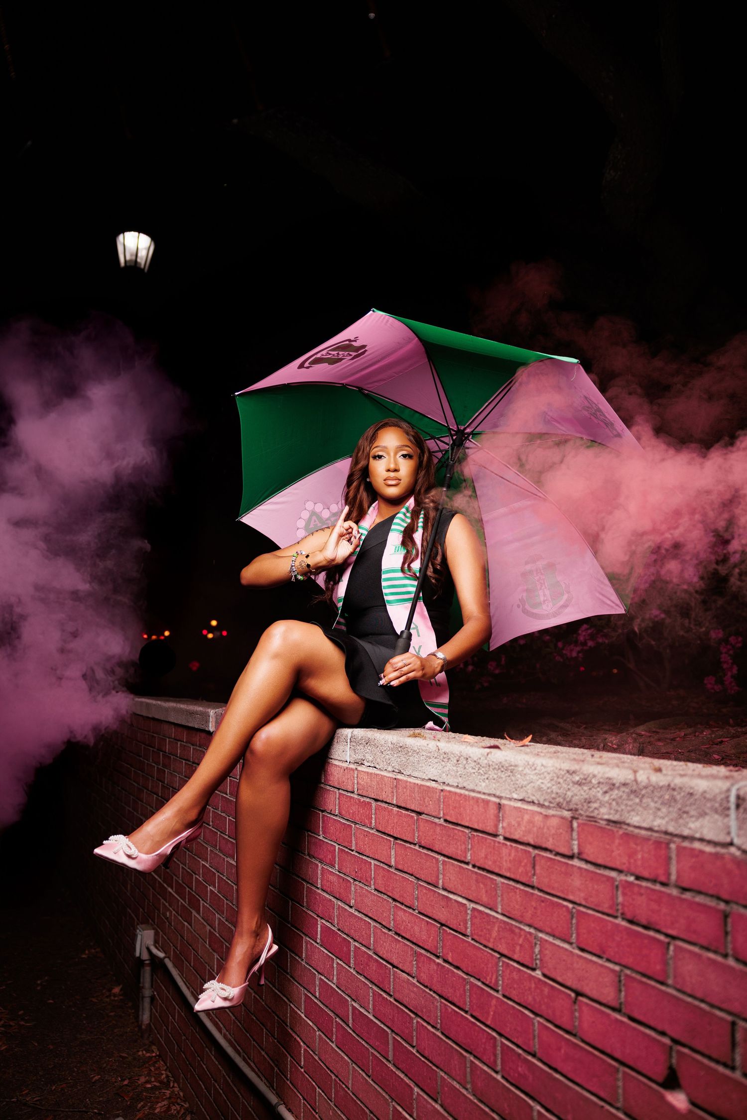 Stylish graduation photoshoot features pink smoke effects and umbrella props on brick wall at night.