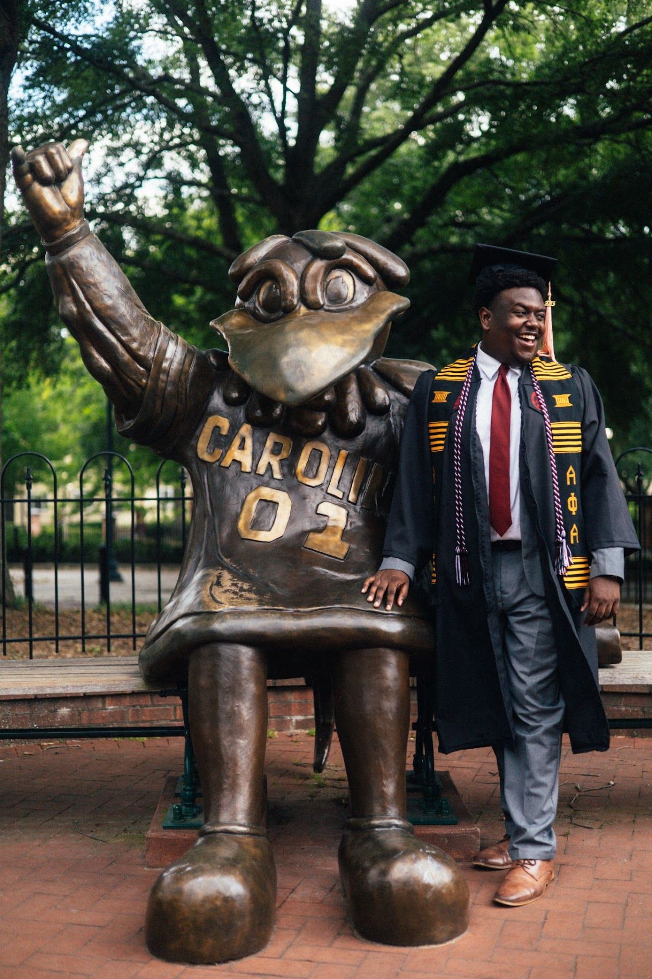 Graduate poses next to bronze mascot statue of Rameses while wearing graduation regalia on USC campus.