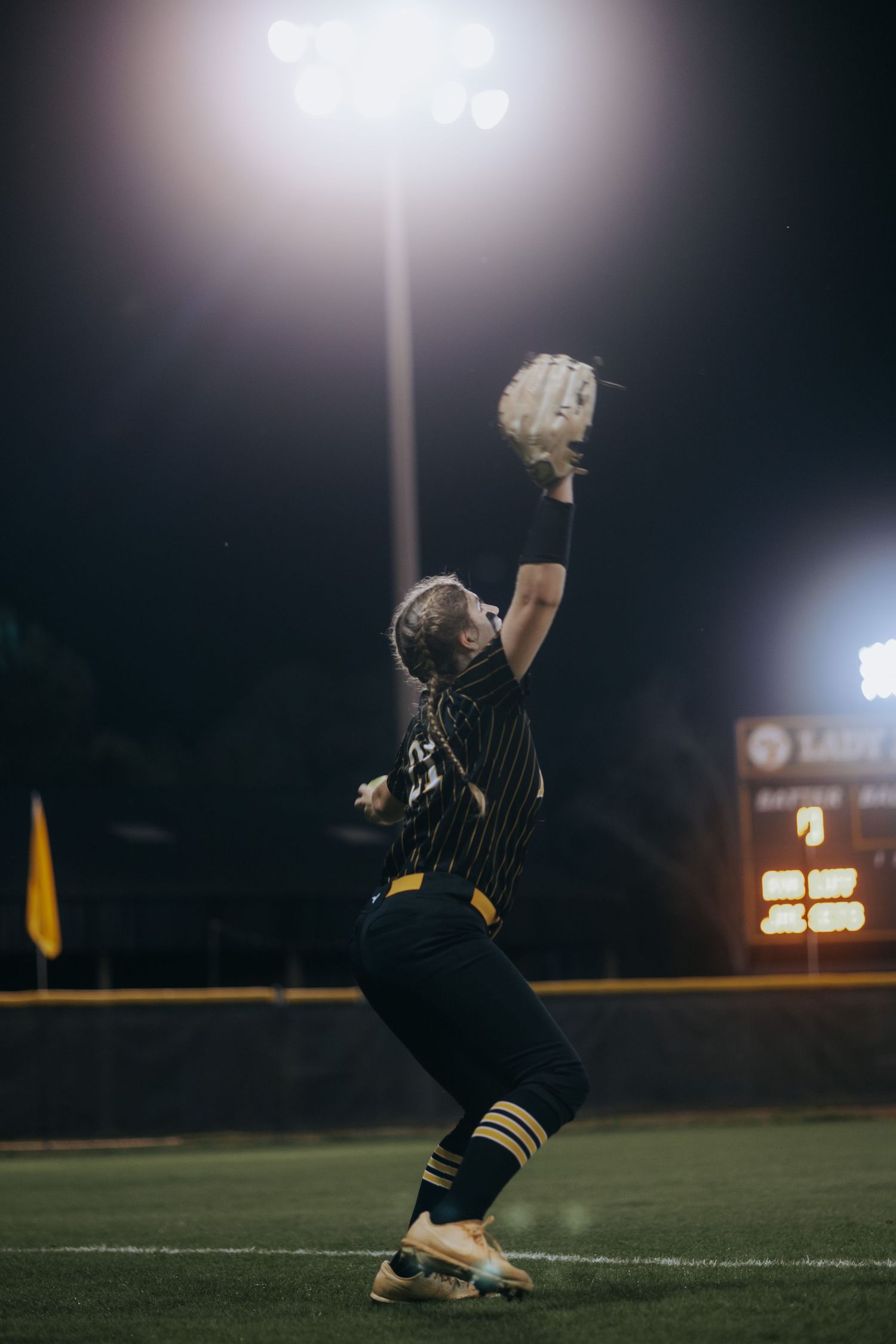 Softball player reaches up to catch ball under bright stadium lights at night.