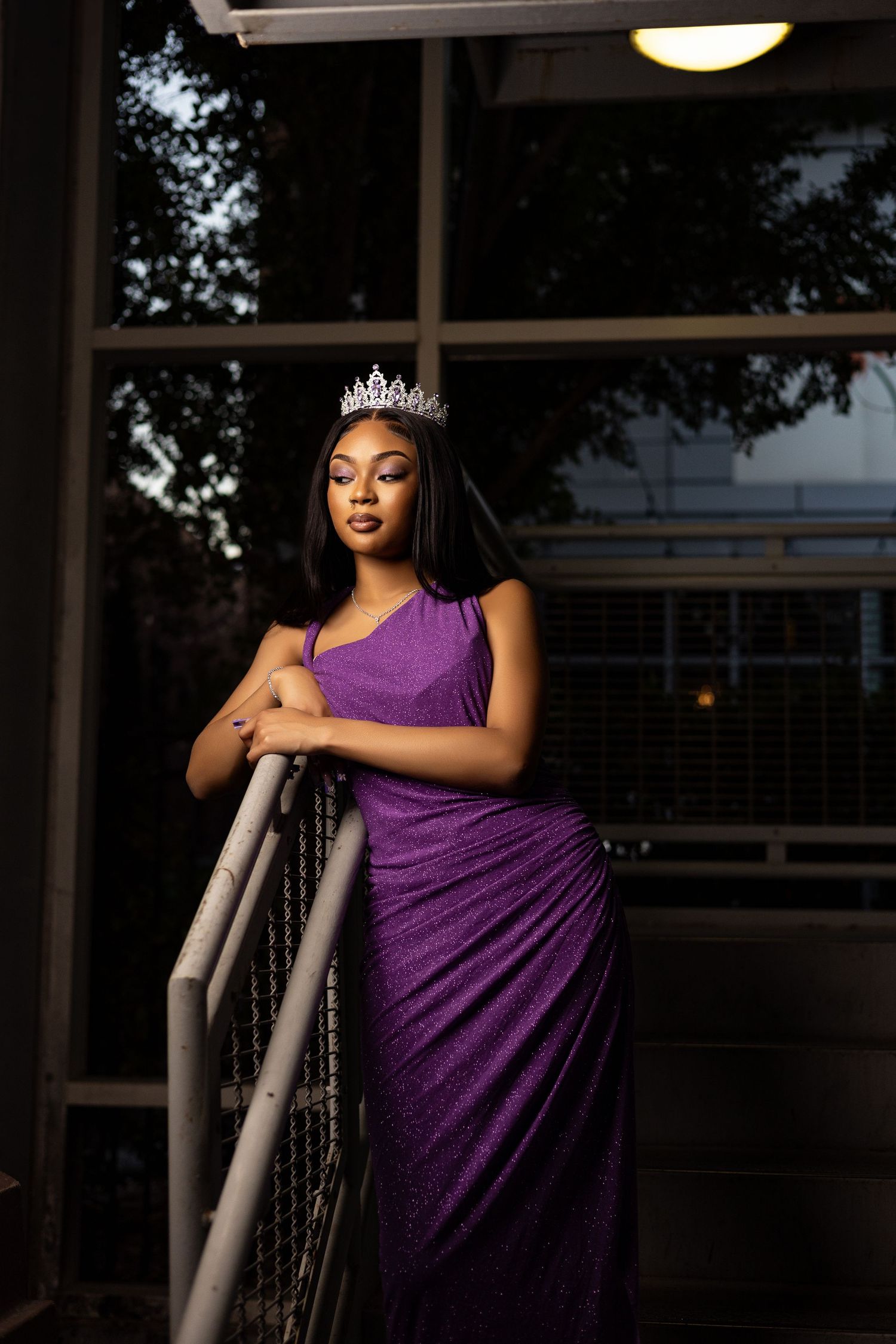 Elegant portrait featuring model in purple gown and tiara by glass windows at dusk.
