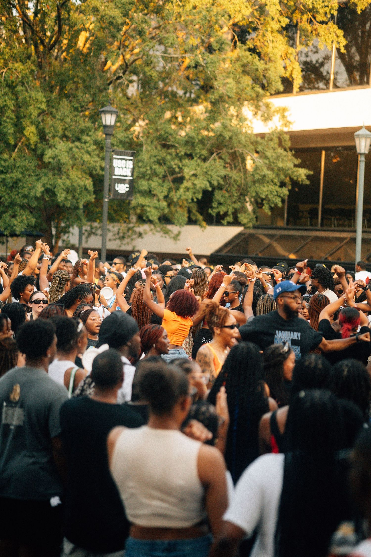 Large energetic crowd of people with raised hands gathering outdoors under yellow autumn trees.USC Hip Hop Wednesday