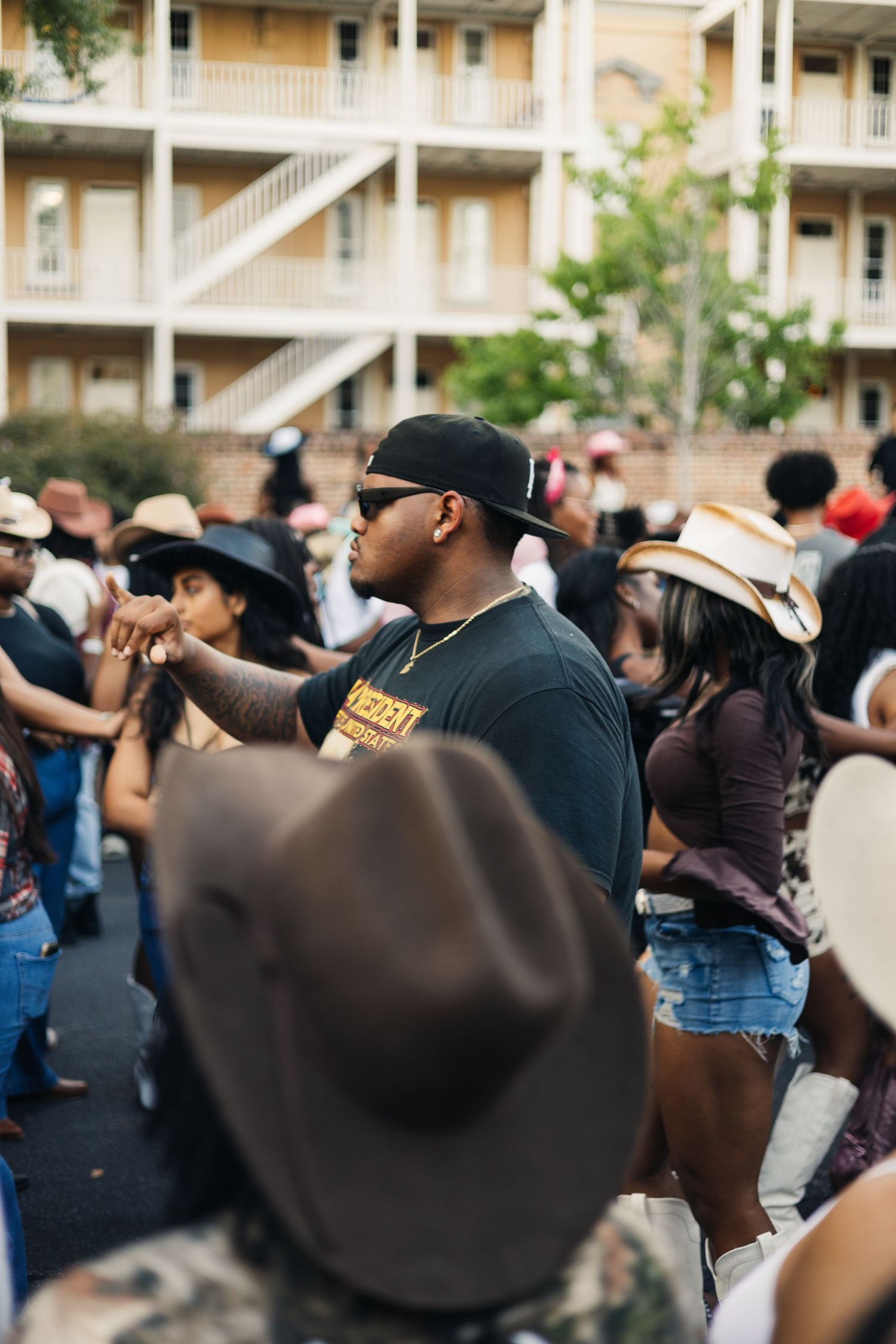 Crowd of people gathered at outdoor event with apartment building in background wearing hats and casual attire.
