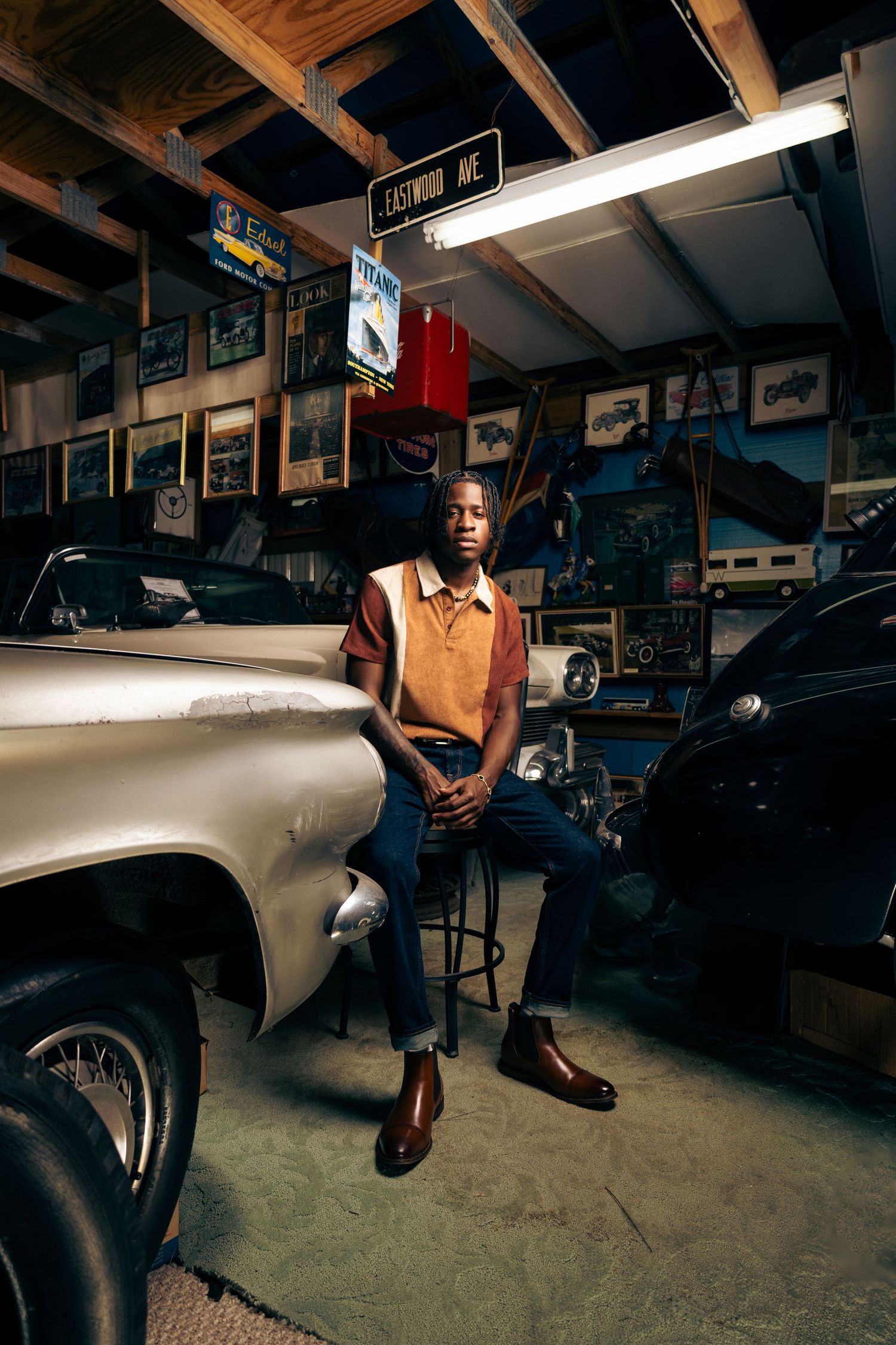 Vintage-style photo of person in brown vest posing beside classic car in garage setting.