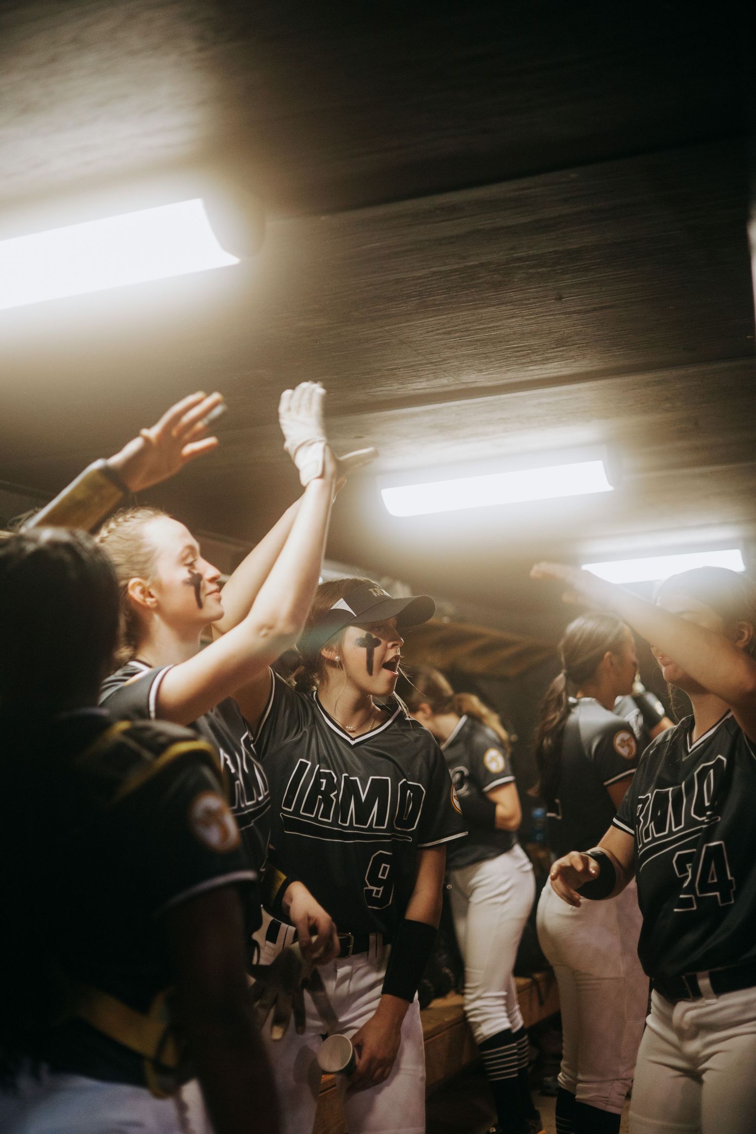 Softball team celebrates with raised arms in locker room under fluorescent lights.