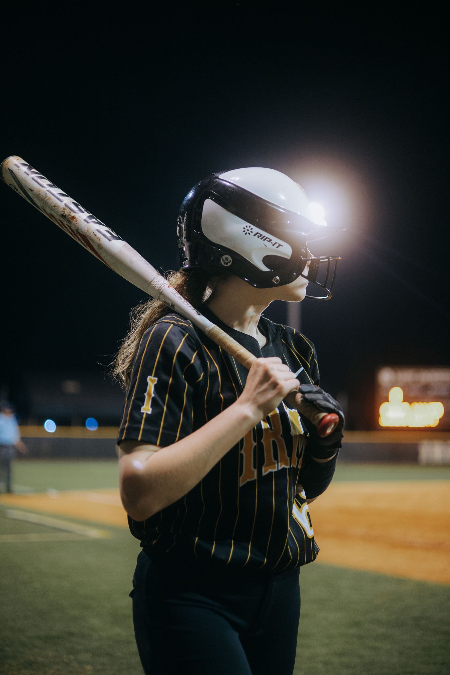 Softball player in pinstriped uniform takes batting stance under stadium lights at night.