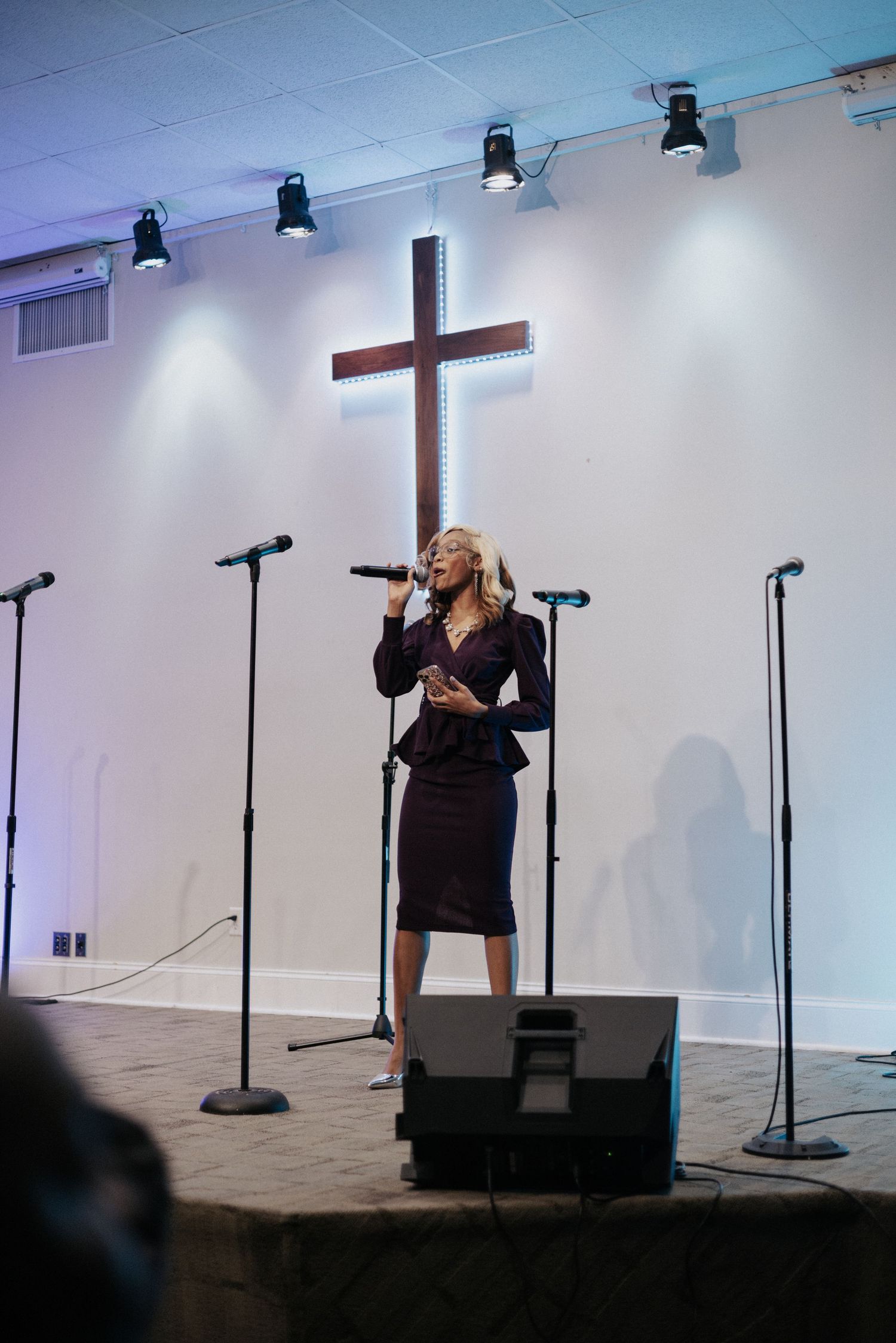A performer sings on stage in front of a large cross with stage lighting in a church setting.