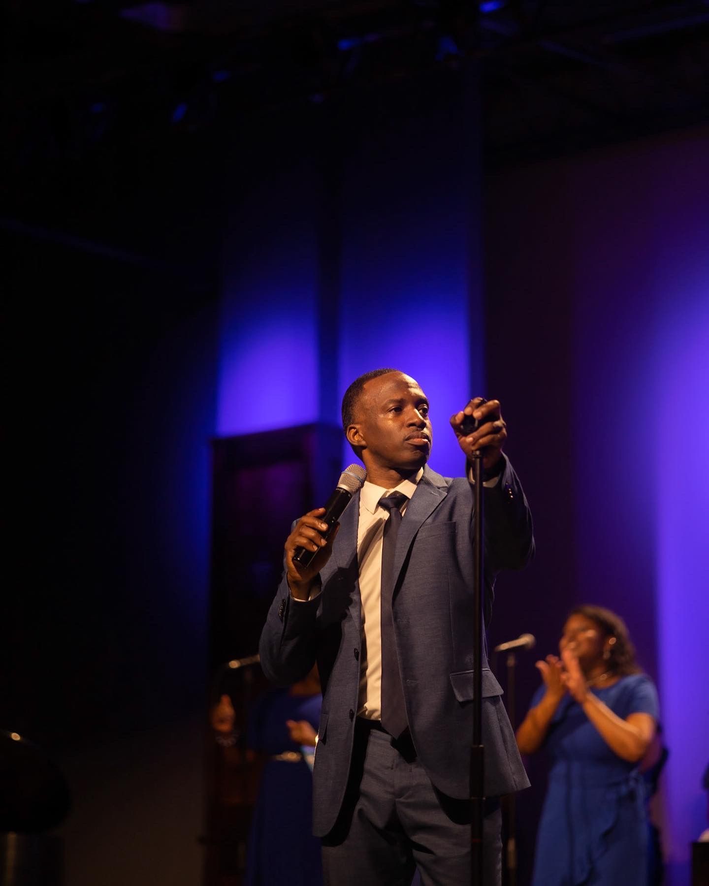 Performer in a dark suit gestures dramatically on stage with dramatic purple lighting.