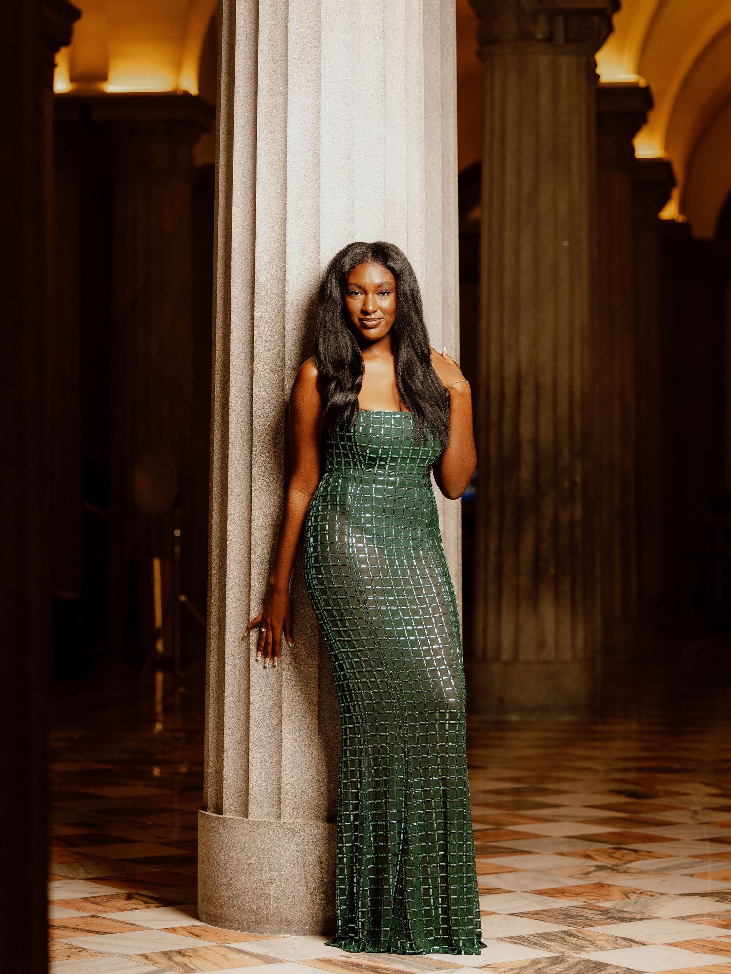 Woman in a green crochet dress poses elegantly against marble columns in an ornate indoor setting.