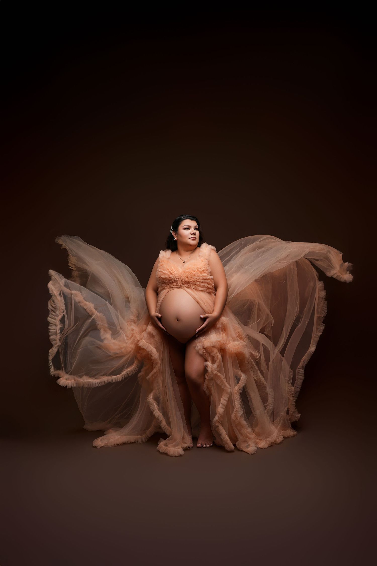 Pregnant woman in flowing peach chiffon maternity gown with fabric billowing dramatically against dark brown studio background.