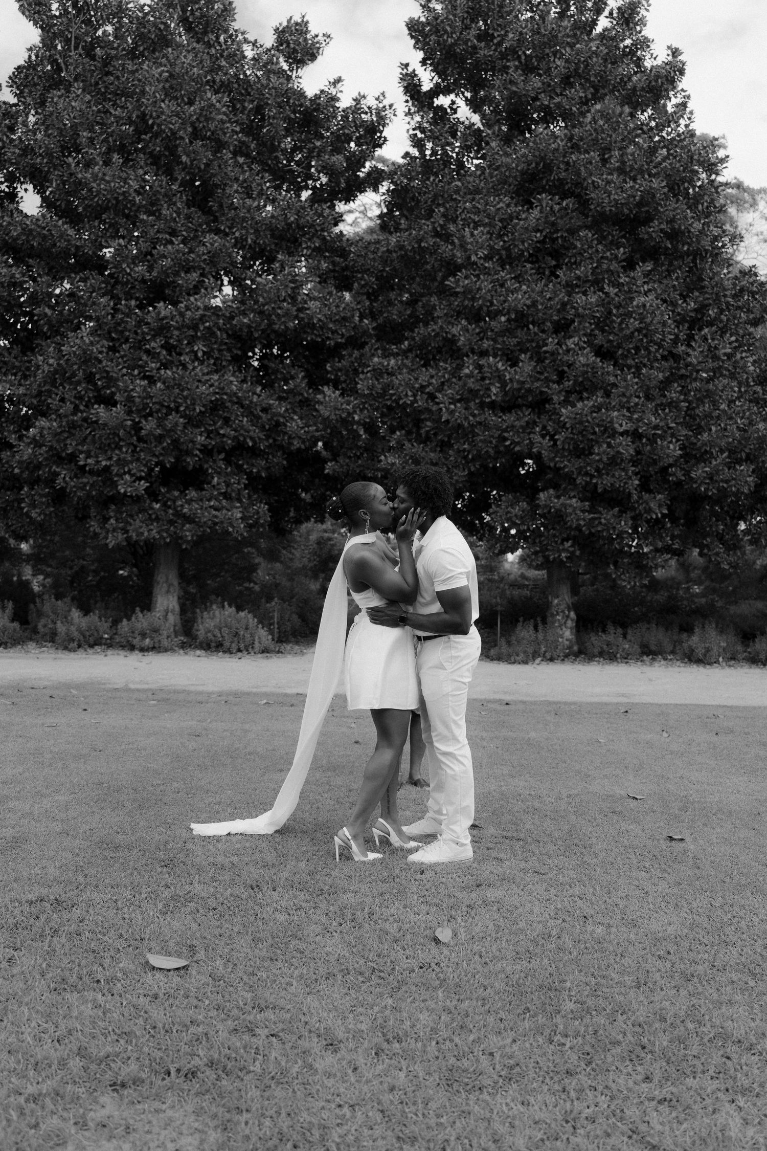 Black and white photo of a couple kissing in wedding attire on a grassy lawn with large trees behind them.