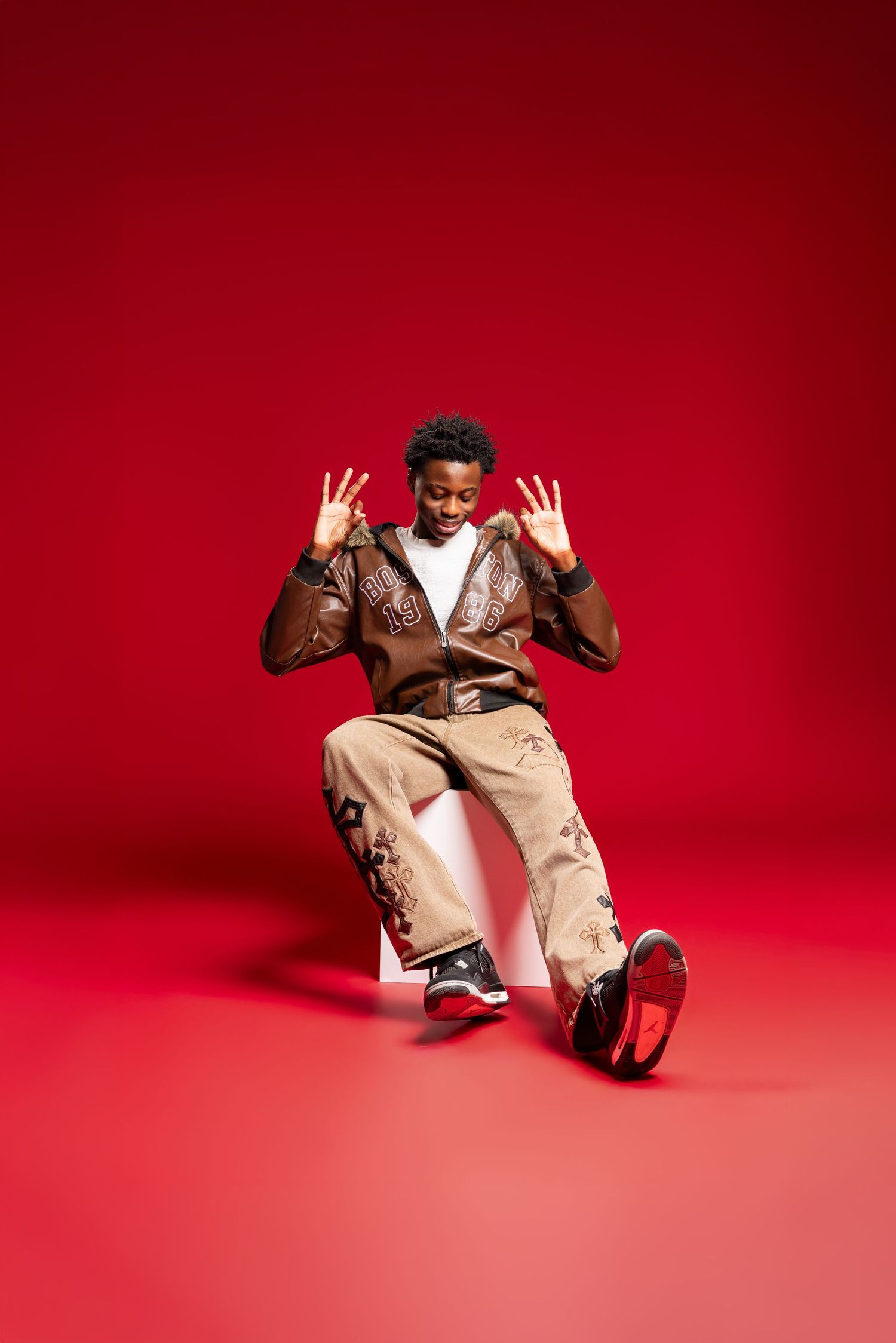 Energetic person in brown leather jacket and khaki pants jumping against vibrant red studio background.