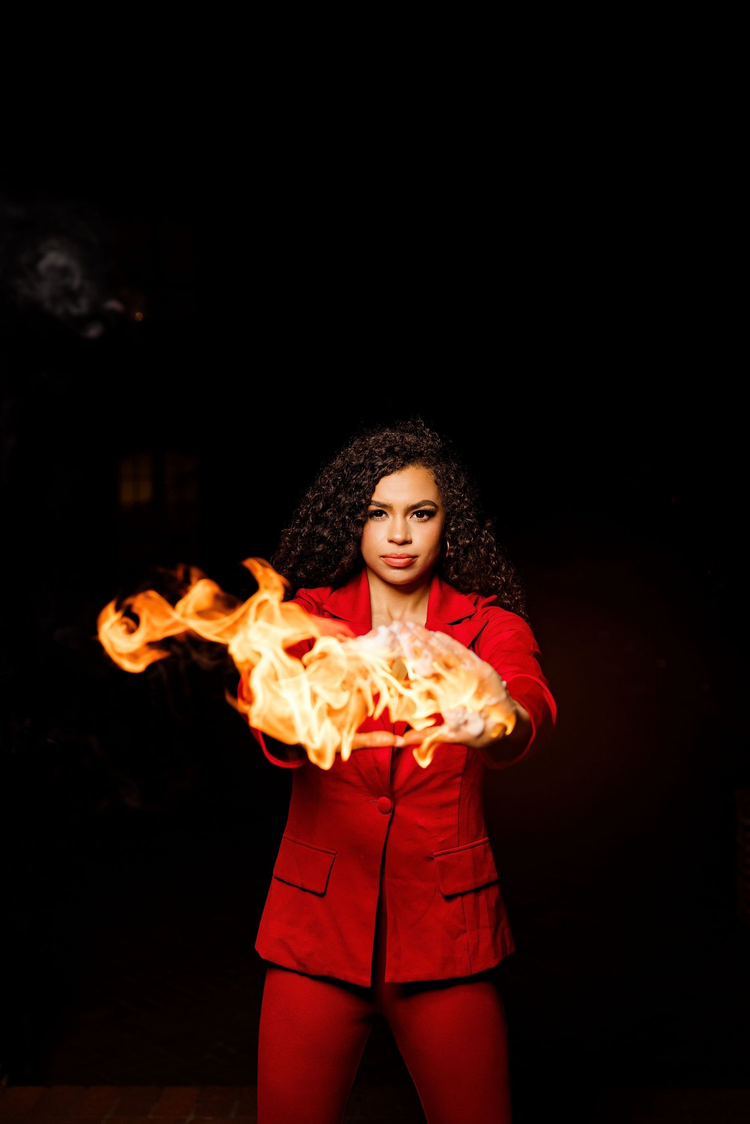 Woman in red suit holding fire in her hands against a dark background.