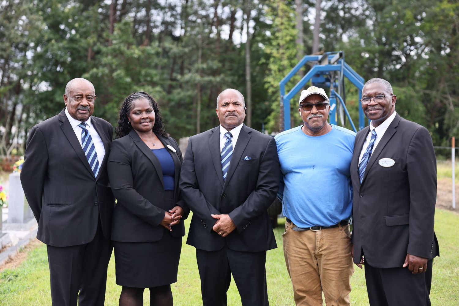 Group of people in formal attire and casual clothing gathered outdoors near pine trees.