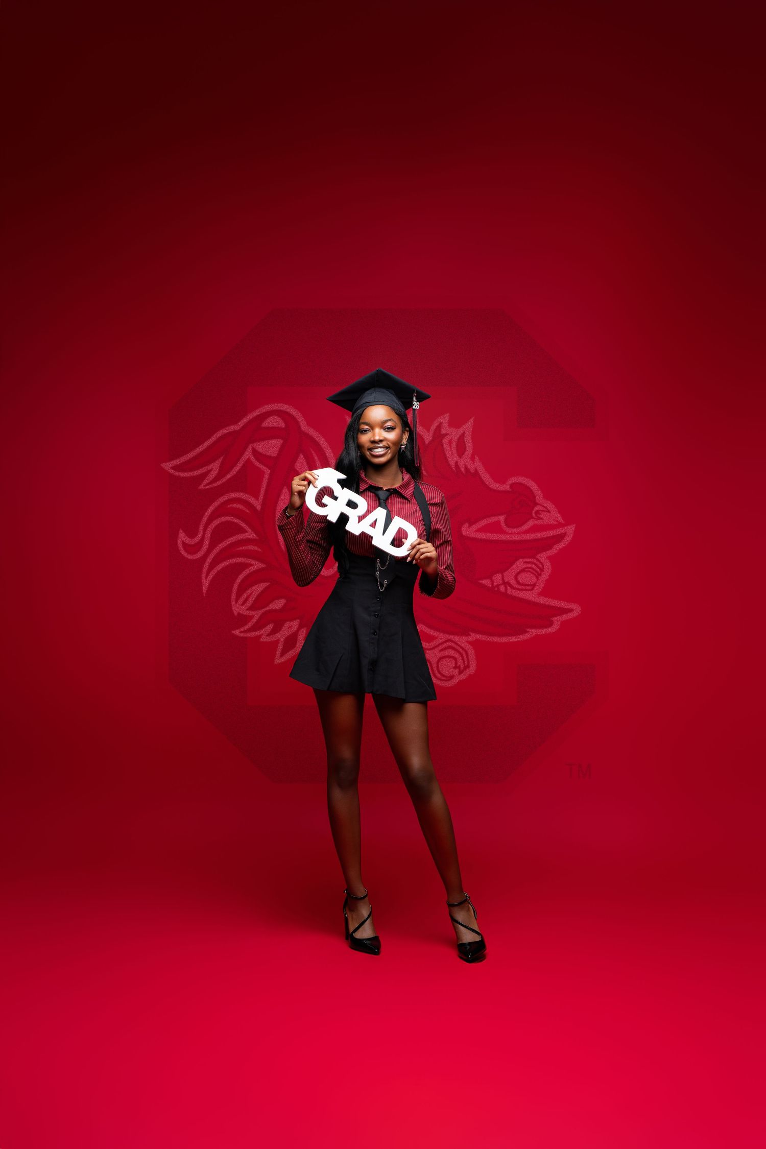 A smiling graduate in cap and black dress holds a GRAD sign against a red background with a university logo.