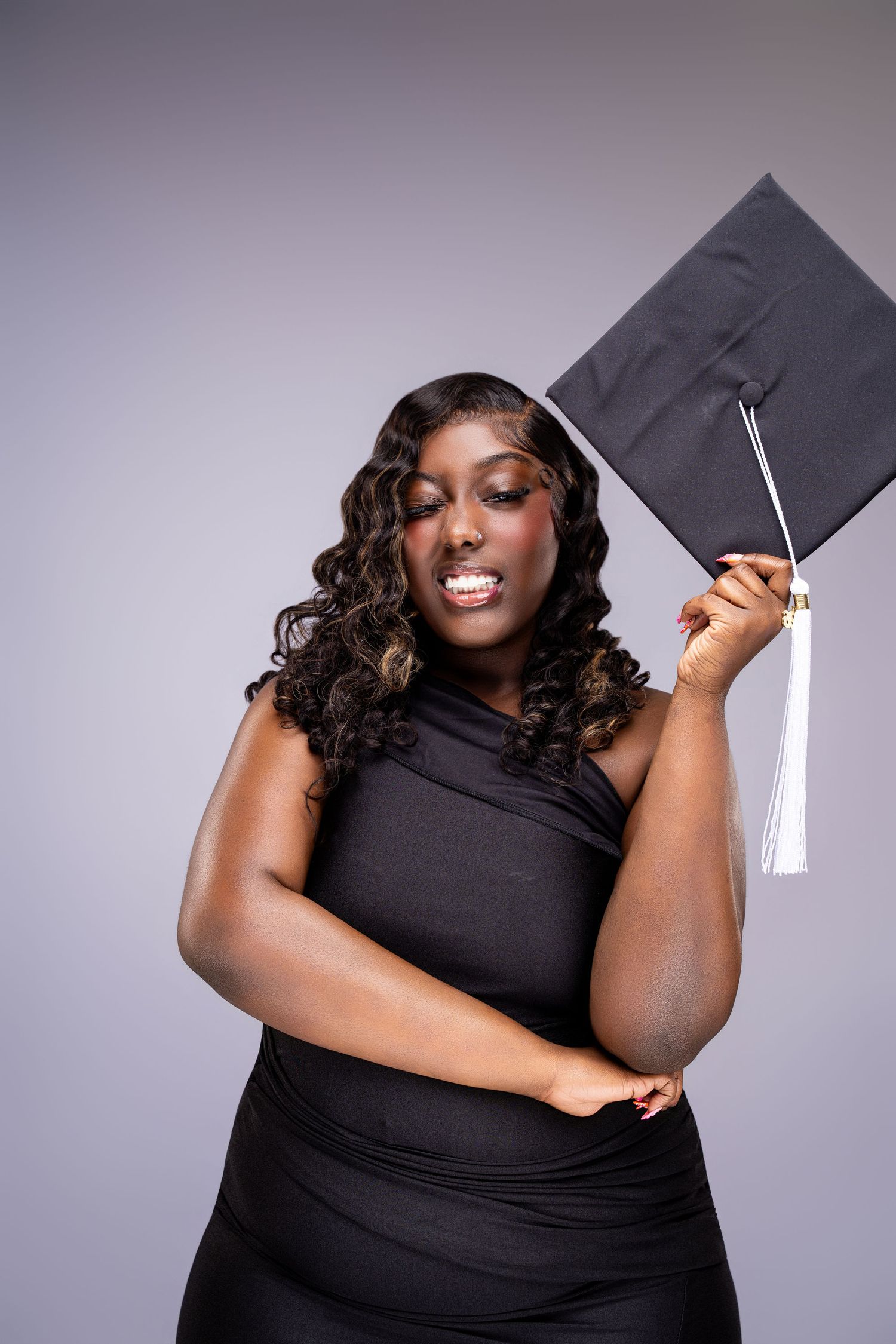 A smiling Black woman in a black dress holds a graduation cap, celebrating her academic achievement.