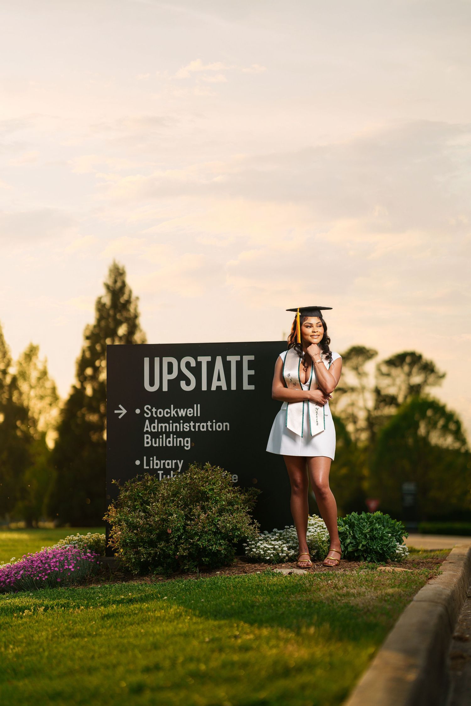 Young woman in white dress and graduation cap poses proudly by USC Upstate campus sign at golden hour.