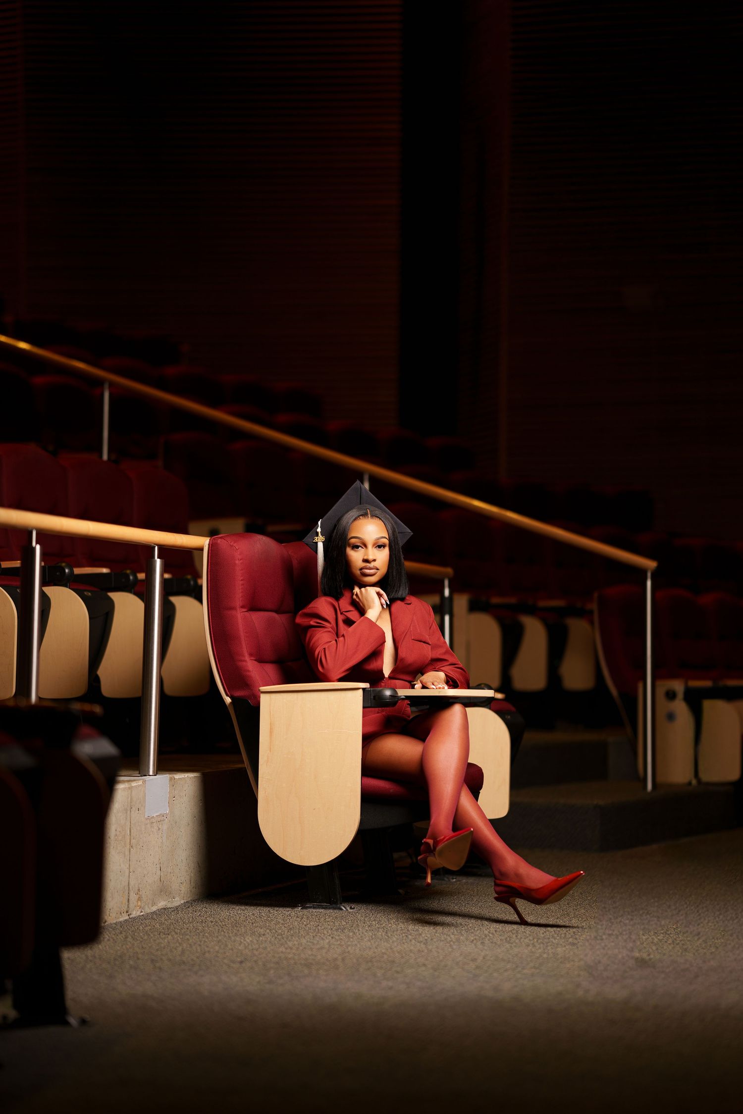 Graduate in red cap and gown sitting elegantly in auditorium chair, posing for graduation portrait photo.