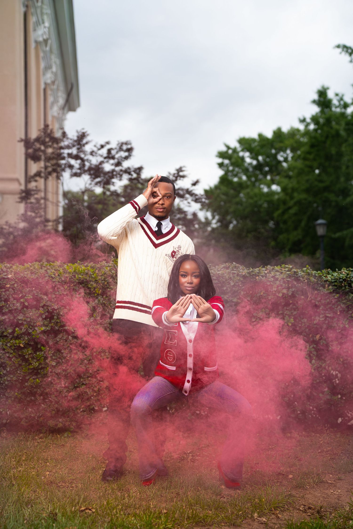 Two people pose dramatically in red smoke outdoors, wearing matching white and red varsity outfits near a building.