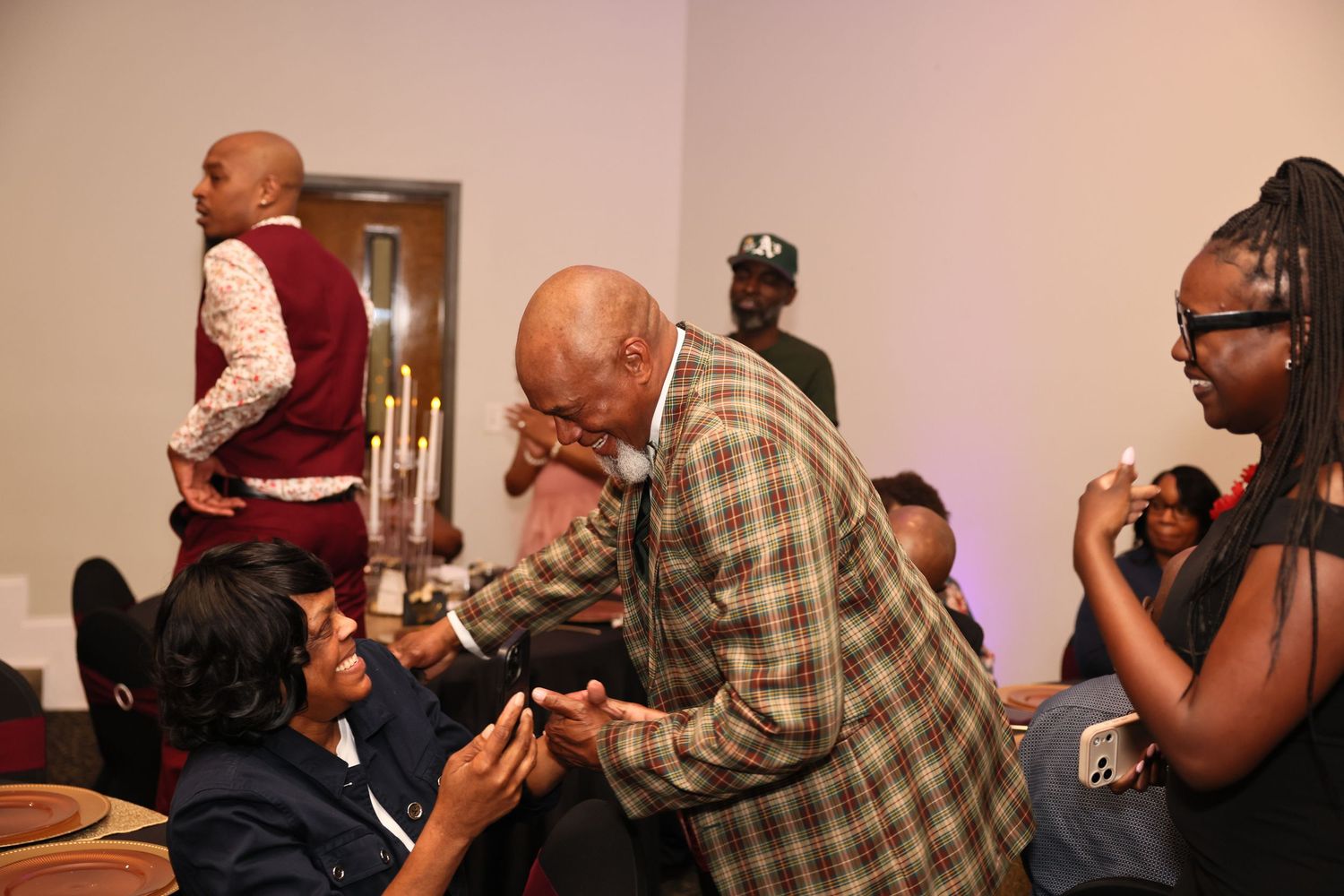 Elderly man in plaid shirt bending down to greet seated woman at a formal indoor gathering with guests.