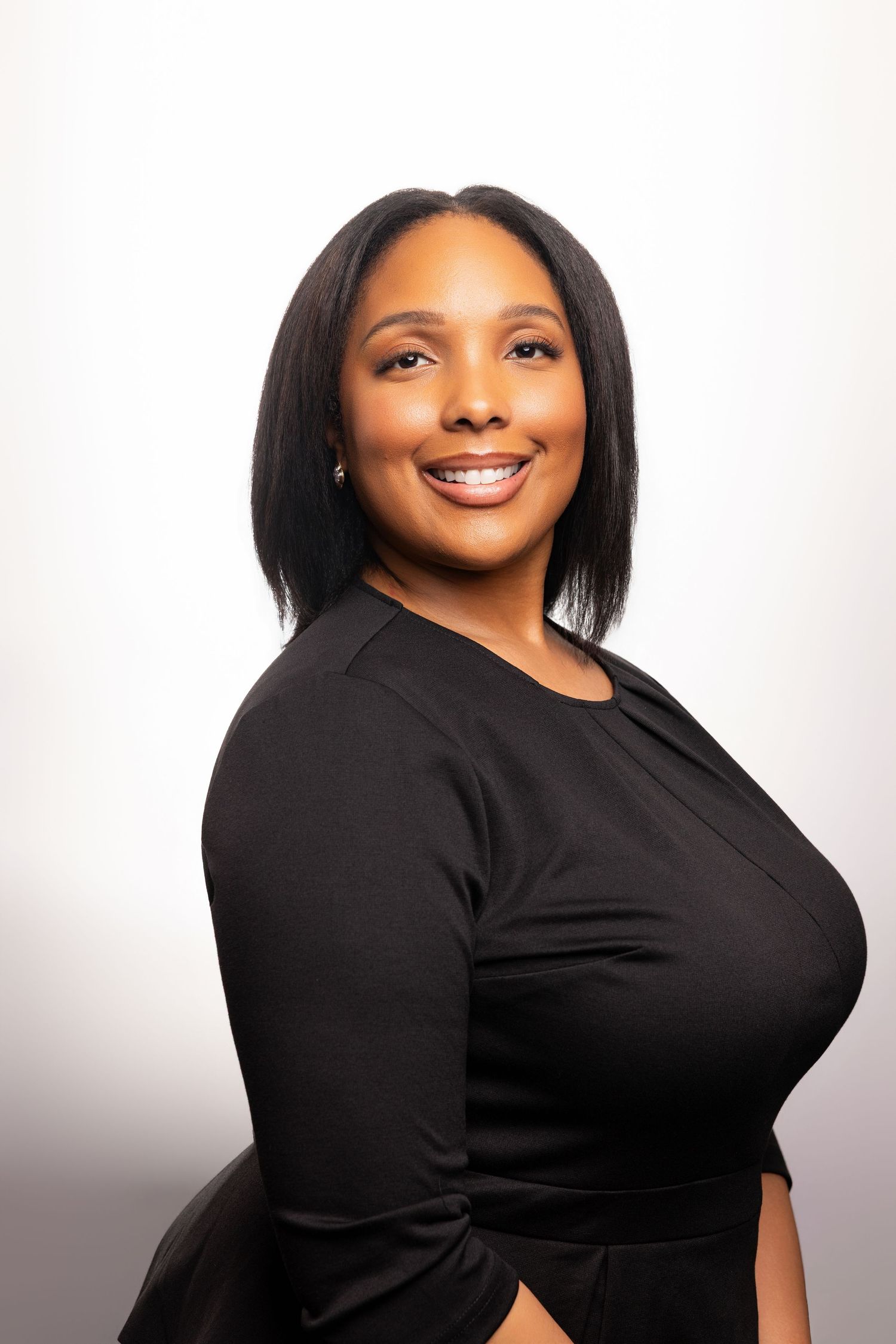 A smiling Black woman with a bob haircut wearing a black dress poses professionally against a light gray background.
