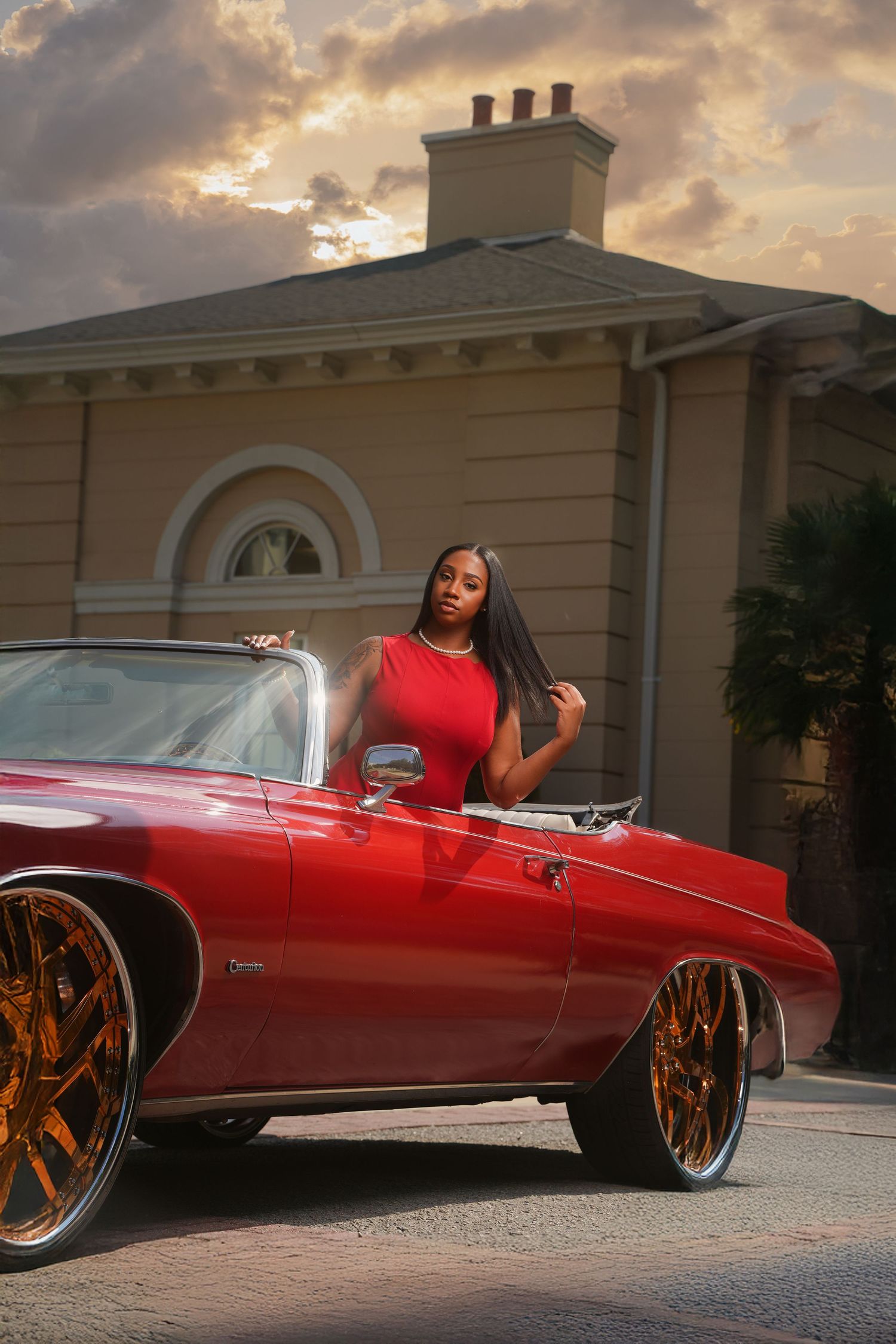 Woman in red dress posing beside a custom red convertible with gold wheels outside an elegant home at sunset.