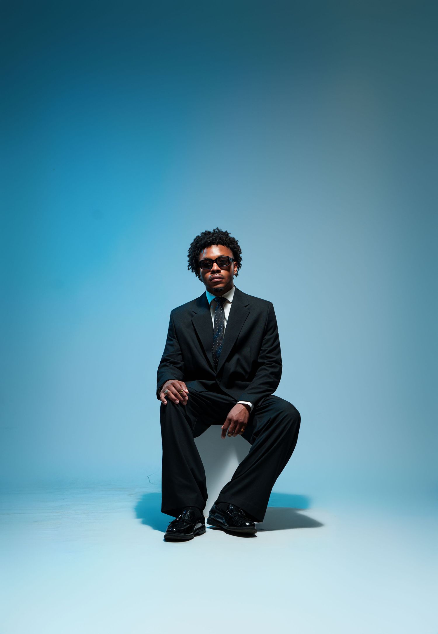 Young man in black suit and sunglasses posing confidently against a blue gradient studio background.
