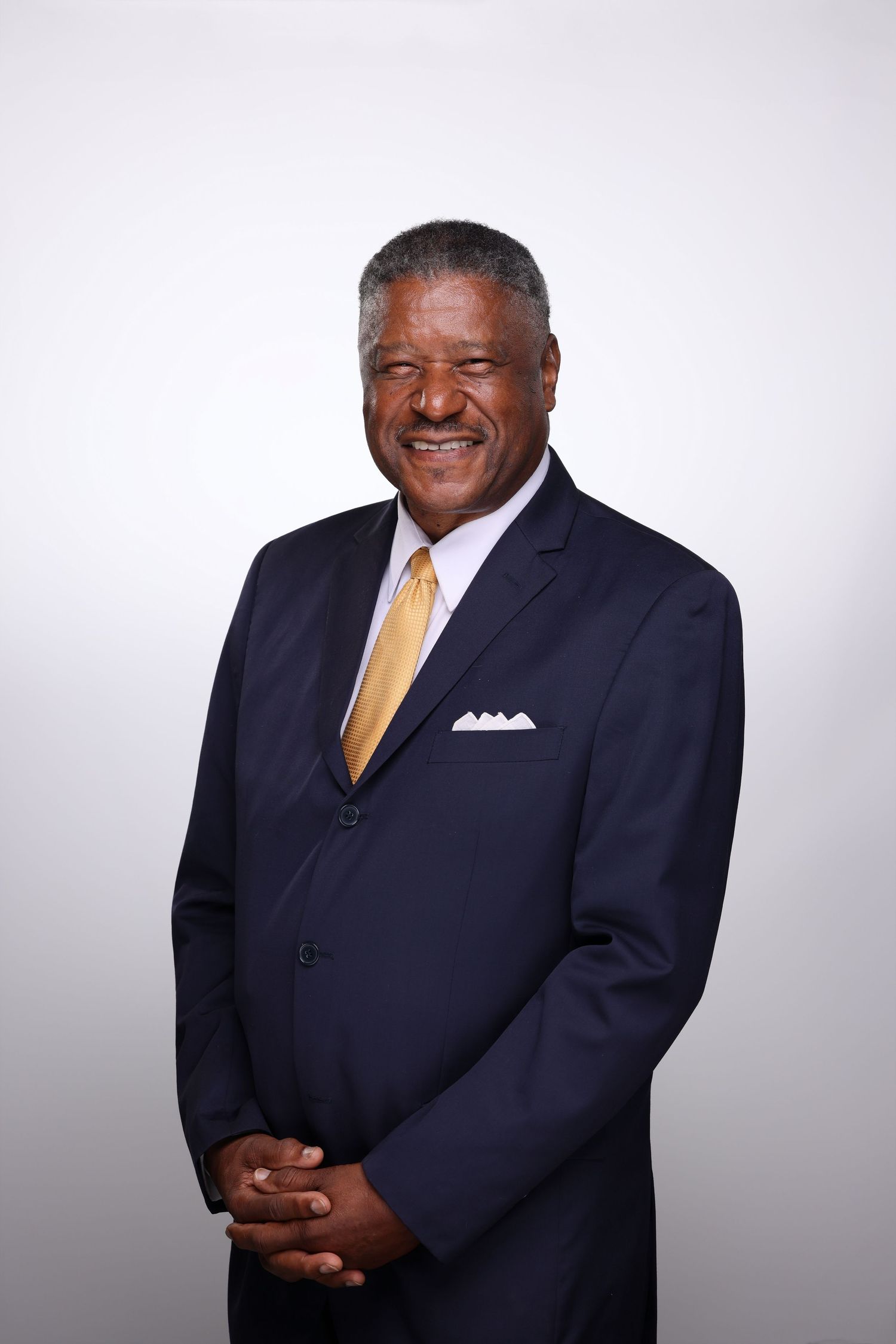 Smiling senior Black man in navy suit, gold tie, and white pocket square poses confidently against gray background.