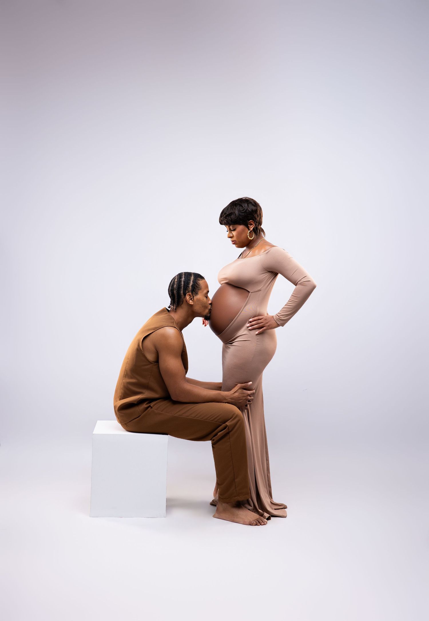 A pregnant woman in a beige gown stands as her partner sits on a white cube and kisses her baby bump in a studio.