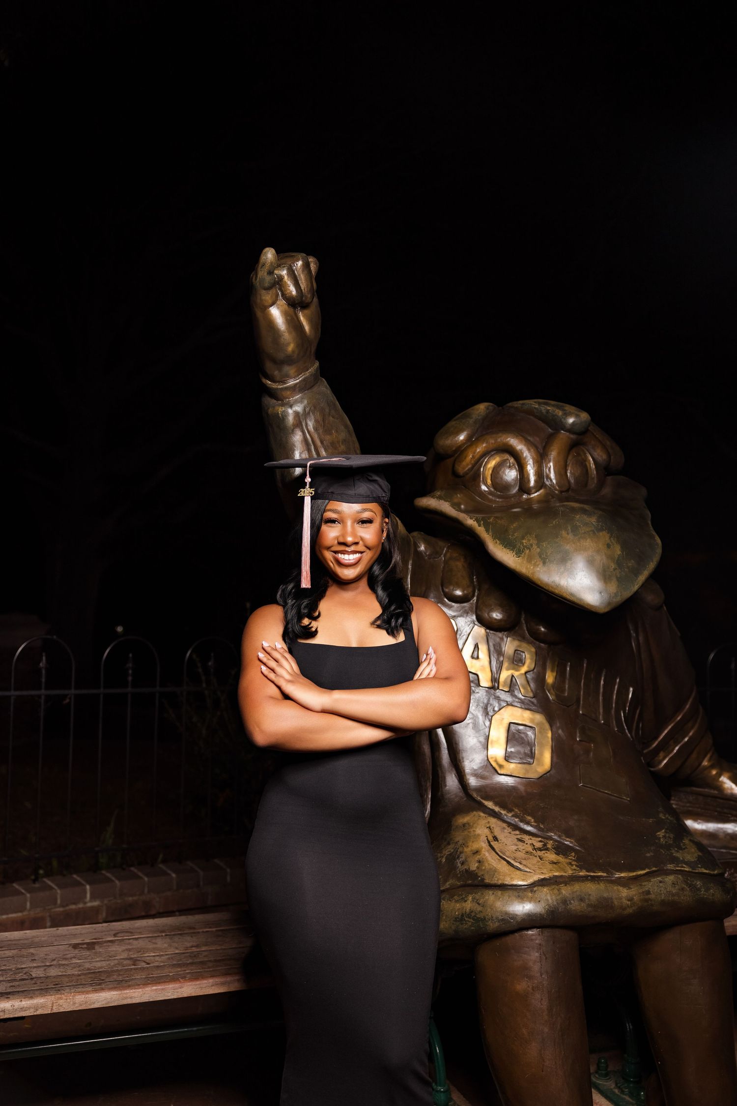 Graduate in cap and gown poses next to bronze mascot statue at night.