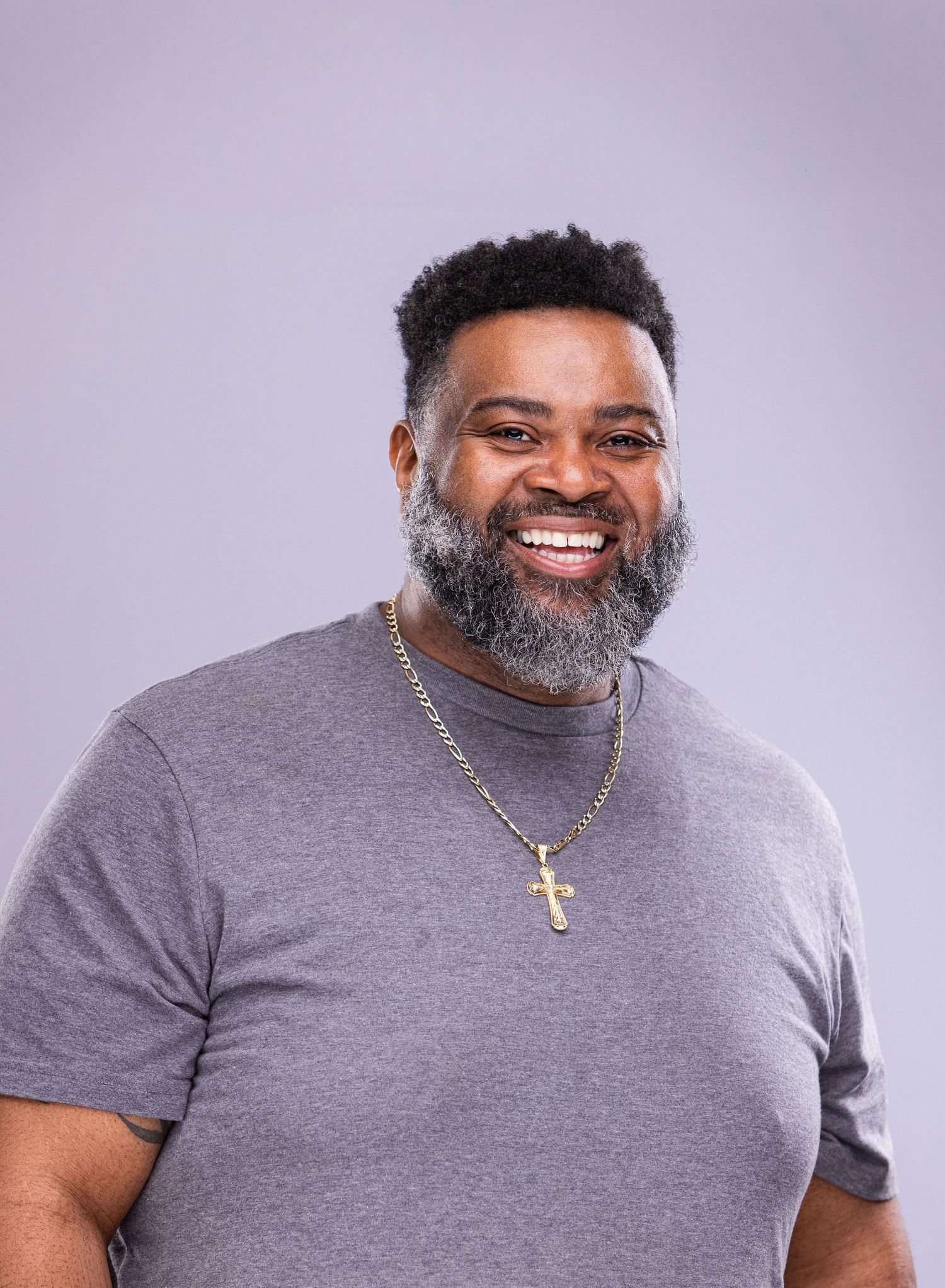 Friendly portrait of person in gray t-shirt with salt and pepper beard against purple background.