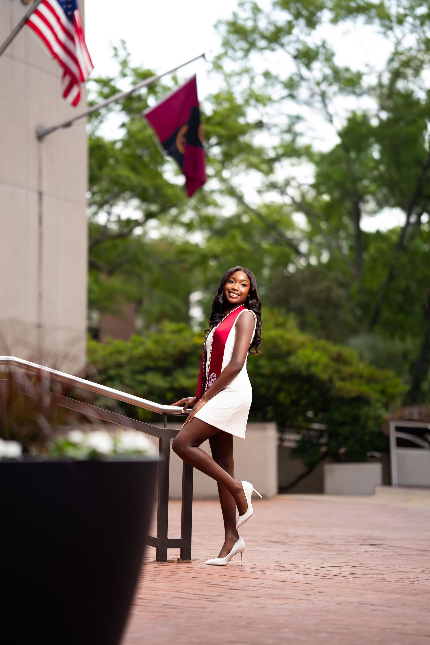 Young Black woman in white and red dress poses elegantly by railing with American flags in background.