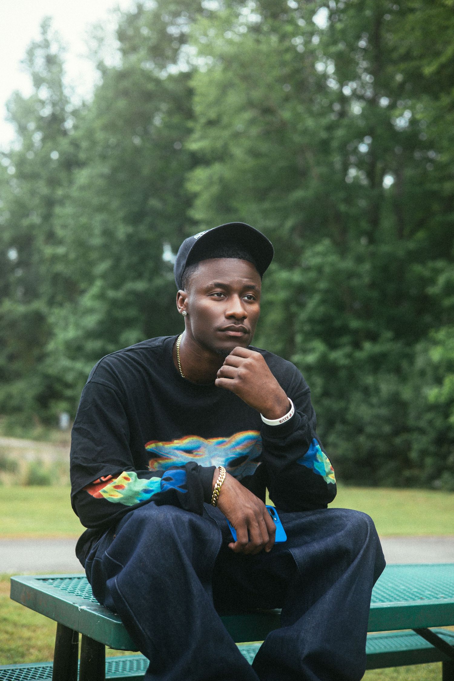 Person wearing black sweater with colorful graphics sits thoughtfully on green picnic table in outdoor setting.