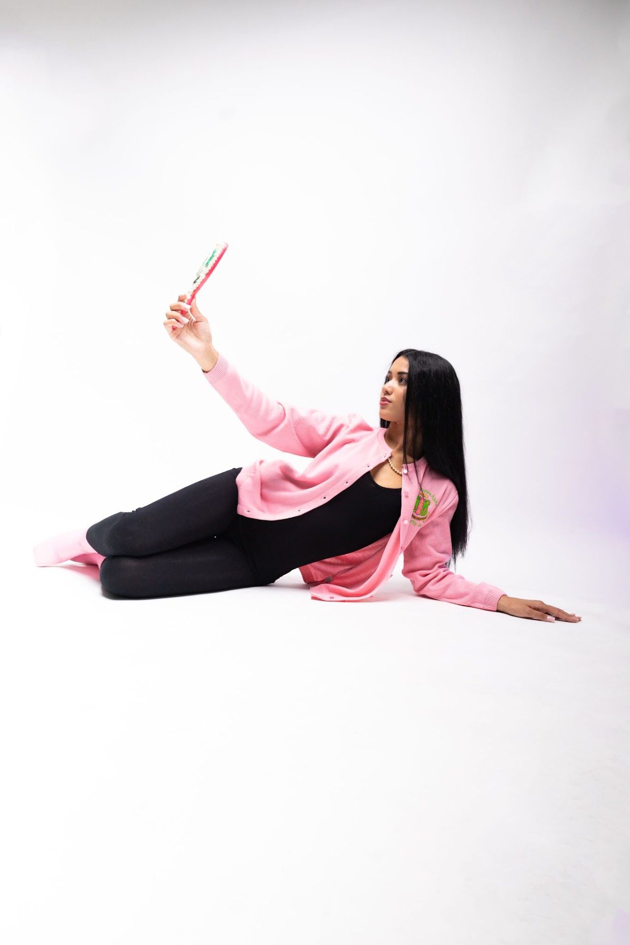 AKA in pink sweater lying on floor holding pink object in artistic pose against white background.