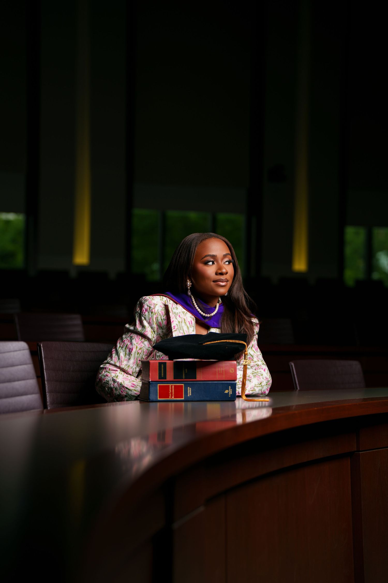 A young Black woman in a floral blazer sits at a conference table with law books, gazing thoughtfully in a formal setting.